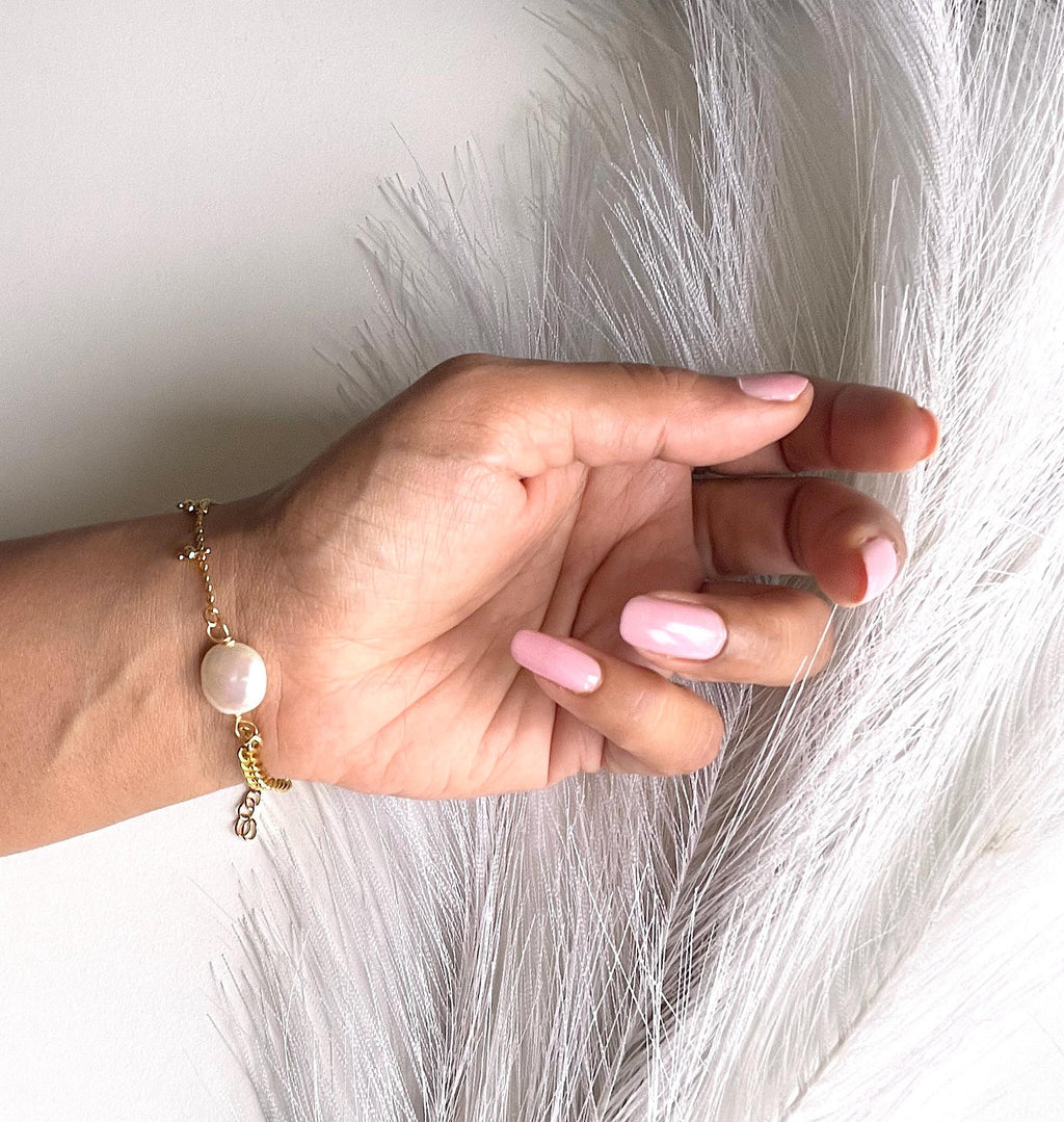 A close-up of a hand with light pink manicured nails wearing a delicate gold chain bracelet with a white oval pearl and a small gold charm, posed against a white background with a fluffy white feather prop.