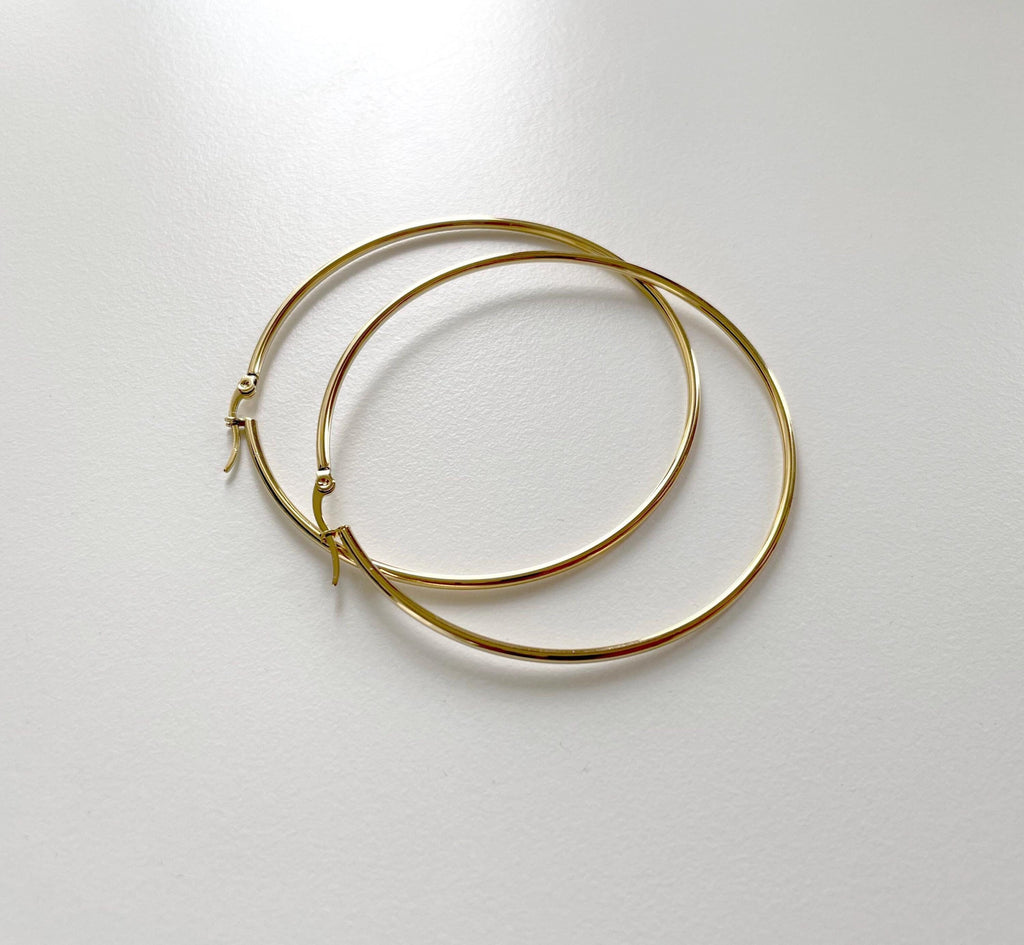 A pair of thin gold hoop earrings with small decorative charms at the closures, displayed on a light gray surface.