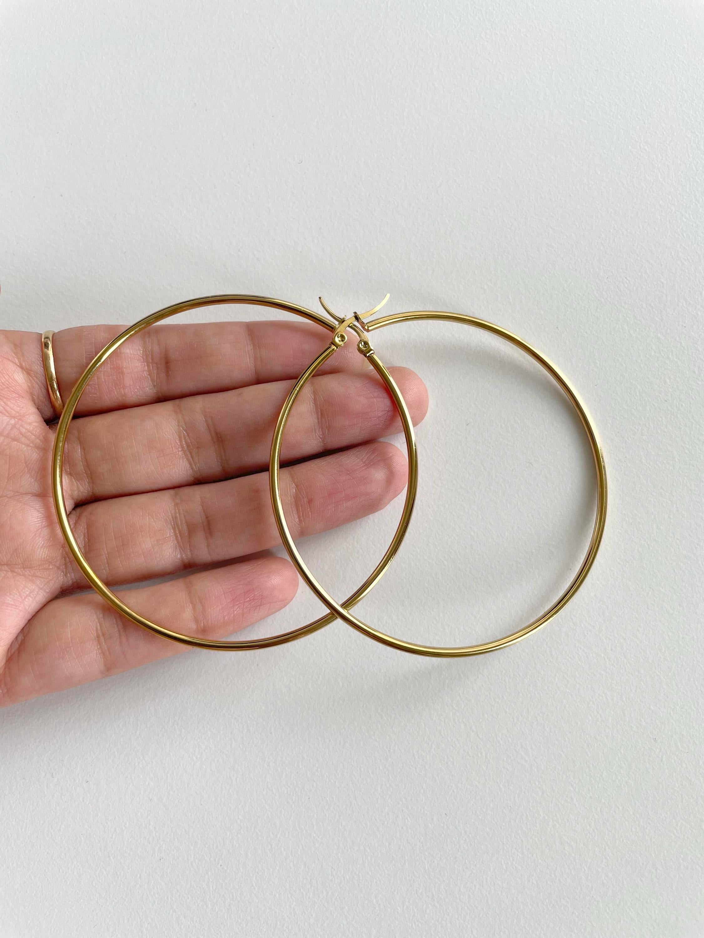 A pair of thin gold hoop earrings held in a person's hand against a plain light background.