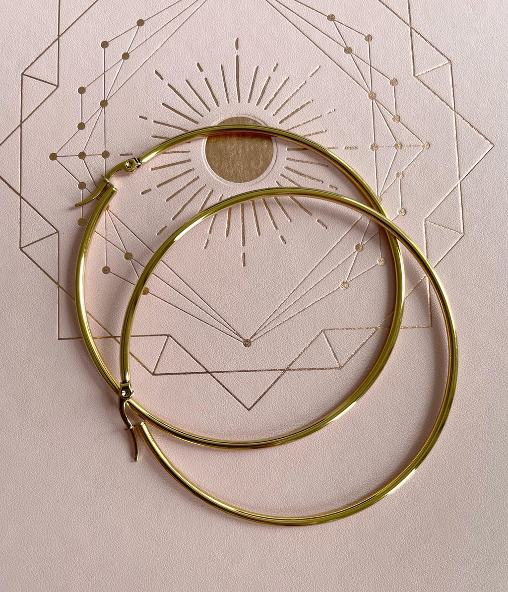 A pair of large, polished gold hoop earrings resting on a light beige textured background with a geometric sun design printed on it.