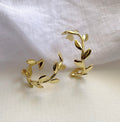A pair of 18K gold plated brass leaf-shaped hoop earrings displayed on a soft white fabric background.

