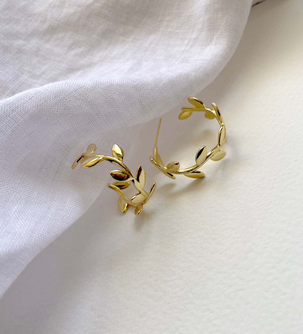 Gold leaf hoop earrings with a delicate vine and leaf design, displayed on a soft white fabric background.

