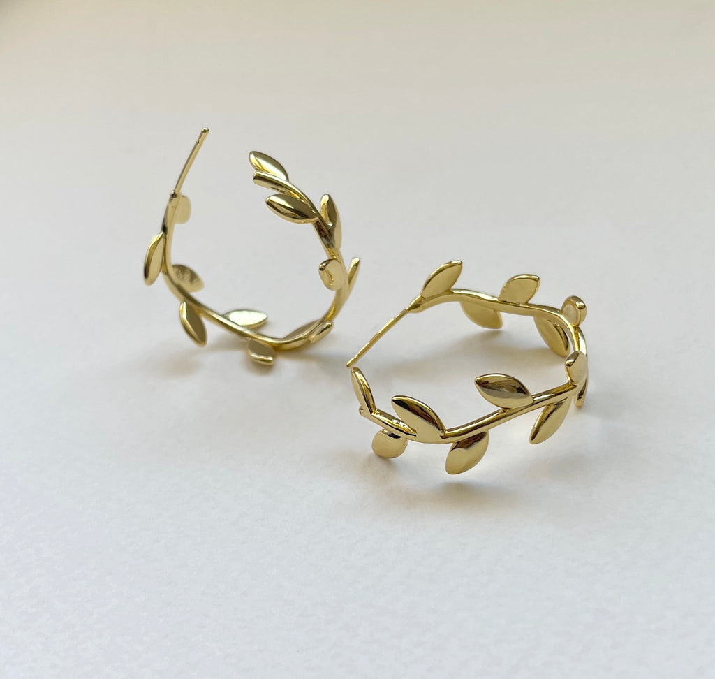 A pair of 18K gold plated leaf wreath hoop earrings on a clean, light off-white background.
