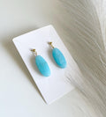 A pair of drop earrings with oval light blue stone pendants and small gold hooks, displayed on a white card against a light neutral background with dried grass.