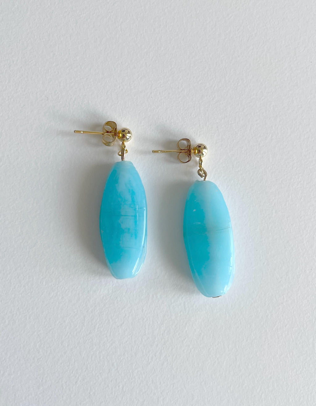 A pair of dangle earrings featuring oval-shaped light blue turquoise stones suspended from gold-tone posts, displayed on a clean, light gray textured background.