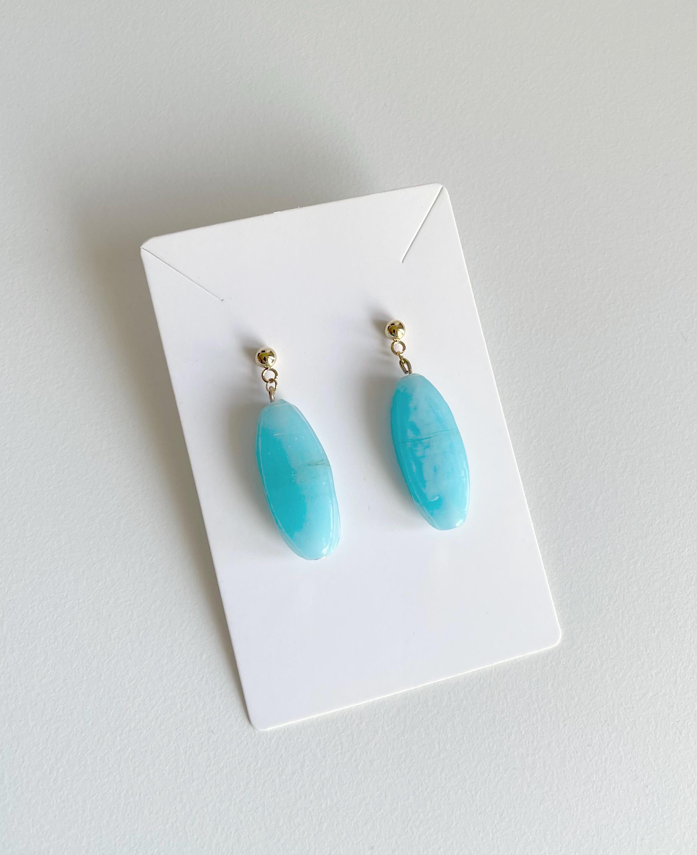 A pair of drop earrings with oval light blue turquoise-like stones and small gold-tone posts, displayed on a white card against a light gray background.