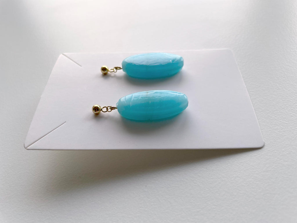 A pair of turquoise-blue oval drop earrings with small gold posts, displayed on a white card against a light gray background.