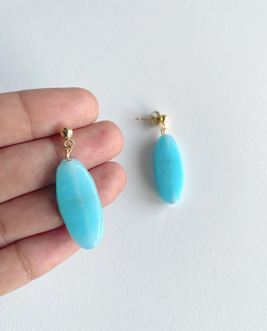 A pair of oval turquoise drop earrings with gold posts, shown with one hanging against a white background and the other held on a person's fingers for scale.