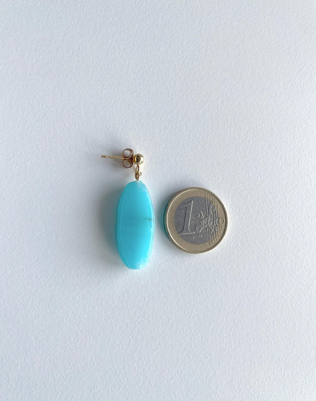A single turquoise oval drop earring with a gold post, shown next to a 1 Euro coin for scale on a white background.