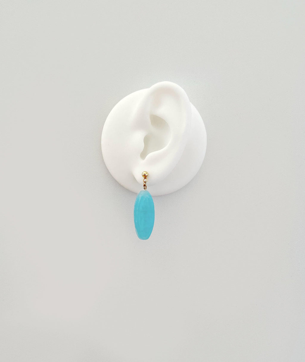 A single drop earring with a small gold-tone stud and a smooth, oval turquoise-blue pendant, displayed on a white ear-shaped stand against a light gray background.