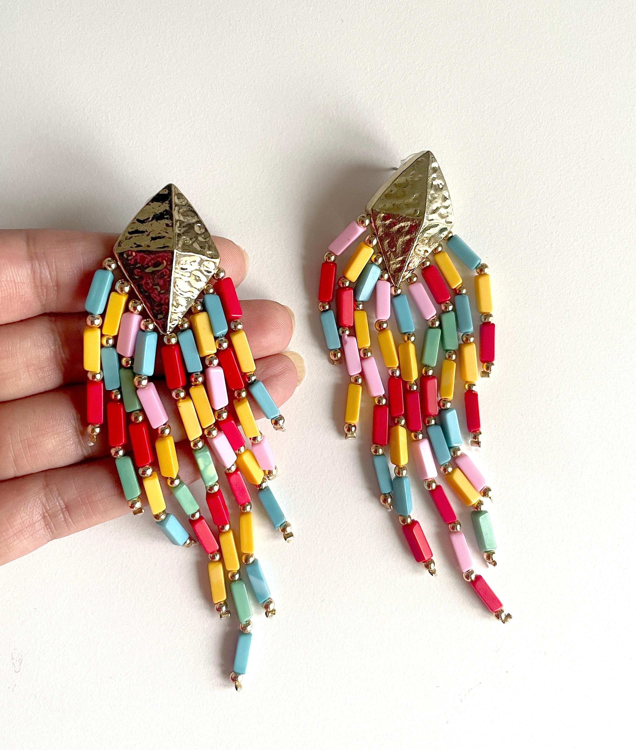 A pair of colorful beaded fringe earrings held in a hand against a plain white background. The earrings feature textured gold-tone metal top pieces and dangling chains of small rectangular beads in red, yellow, light blue, pink, and mint green.