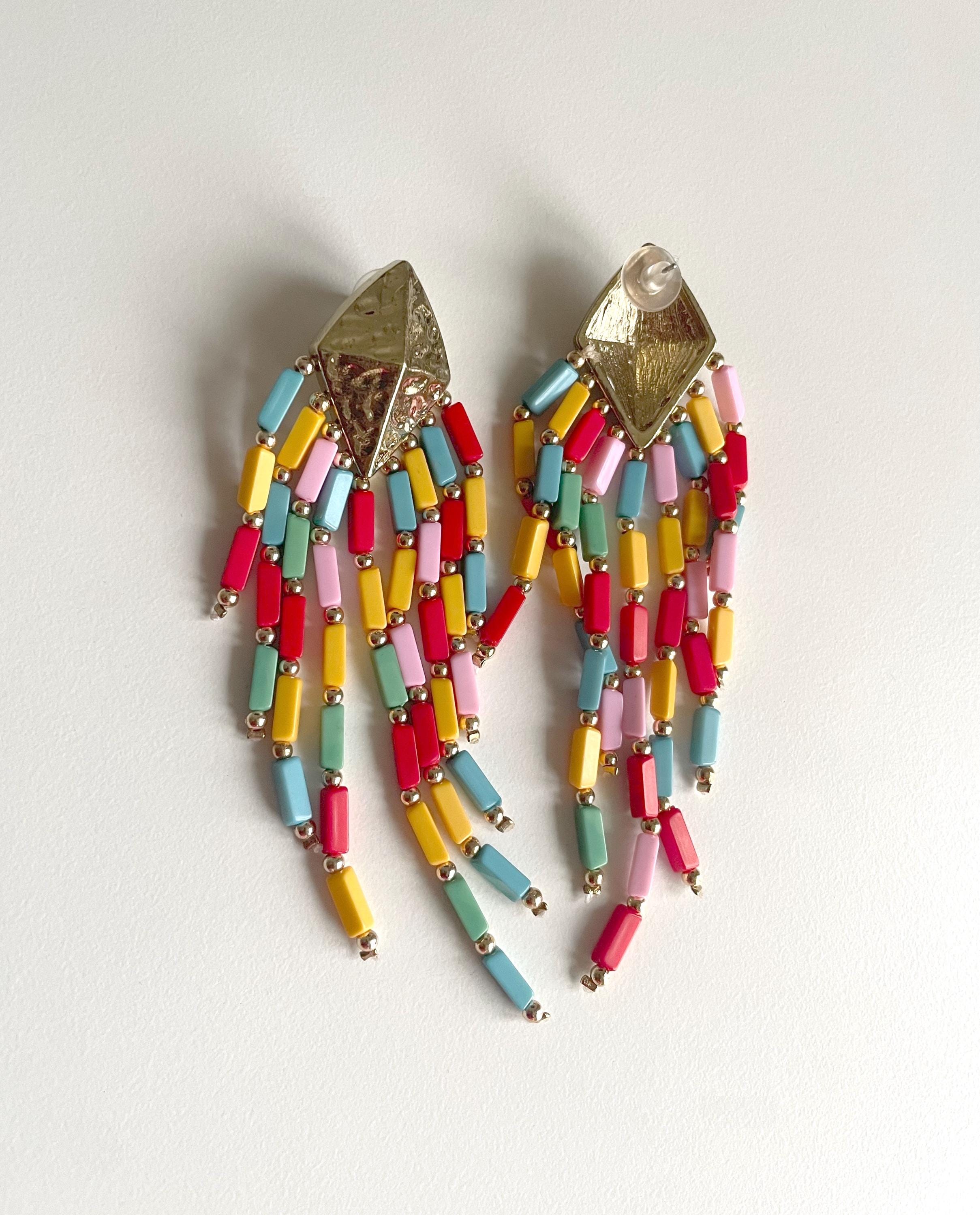 A pair of gold-tone statement earrings with dangling fringe made of colorful cylindrical beads in pink, yellow, light blue, and mint green, photographed on a plain off-white background.
