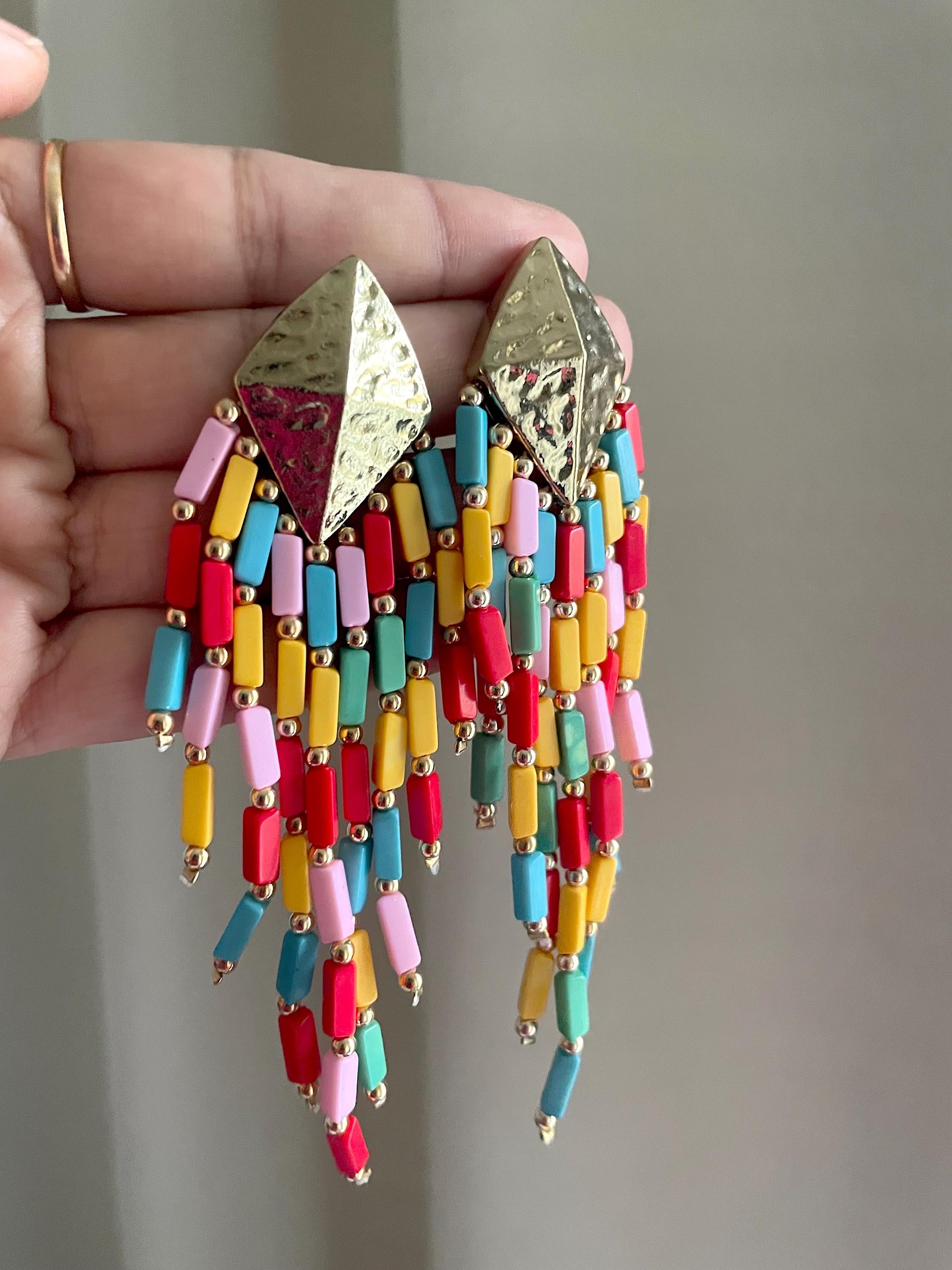 A pair of colorful beaded dangle earrings held in a hand. The earrings feature textured gold-tone triangular studs and long, dangling cylindrical beads in red, pink, yellow, light blue, and turquoise, accented with small gold-tone spacers.