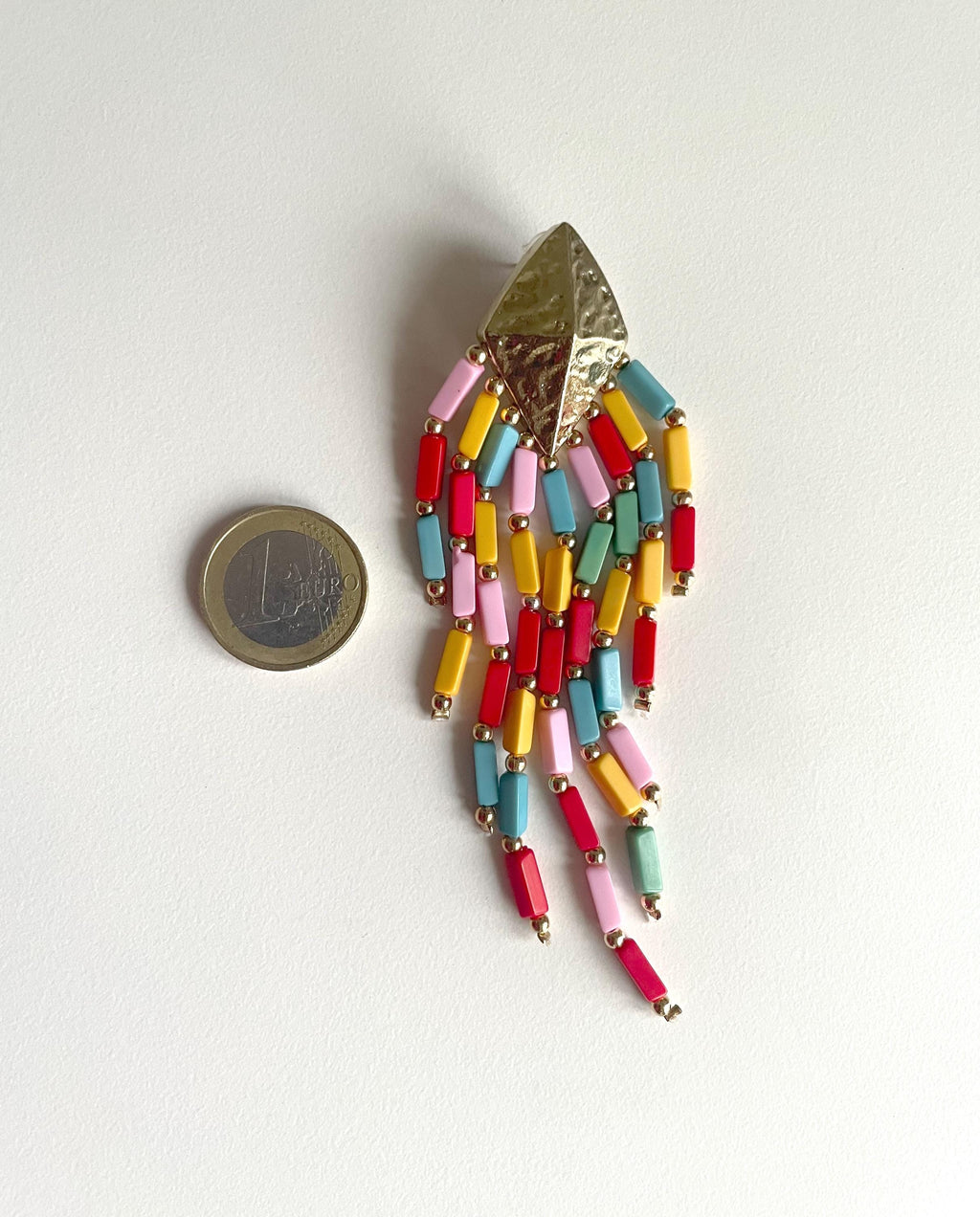 A colorful beaded fringe brooch with a textured gold-tone triangular top, displayed next to a 1 Euro coin for scale on a plain white background.