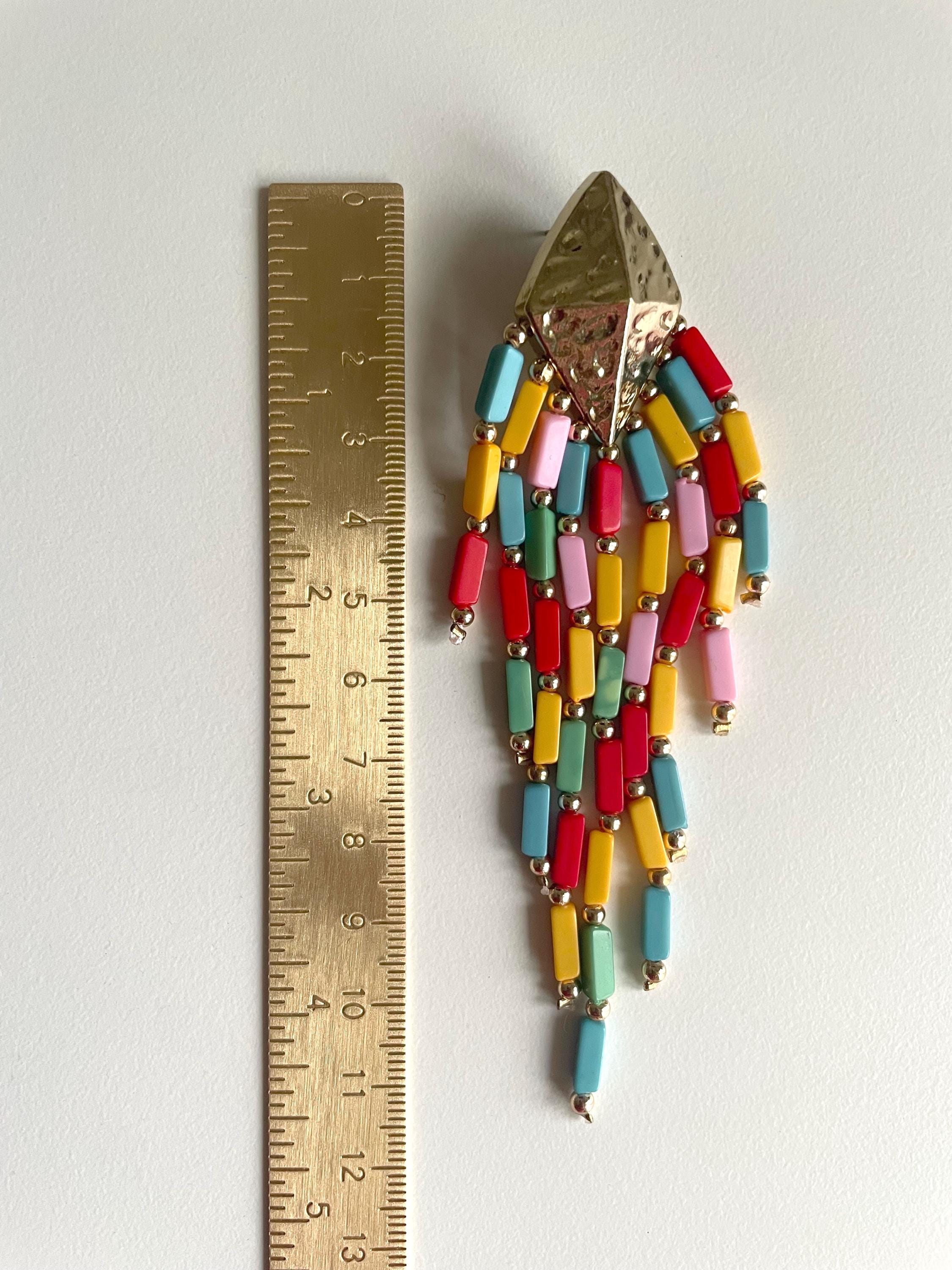 A colorful beaded fringe earring with a gold-toned textured top, shown next to a brass ruler for scale on a plain white background.