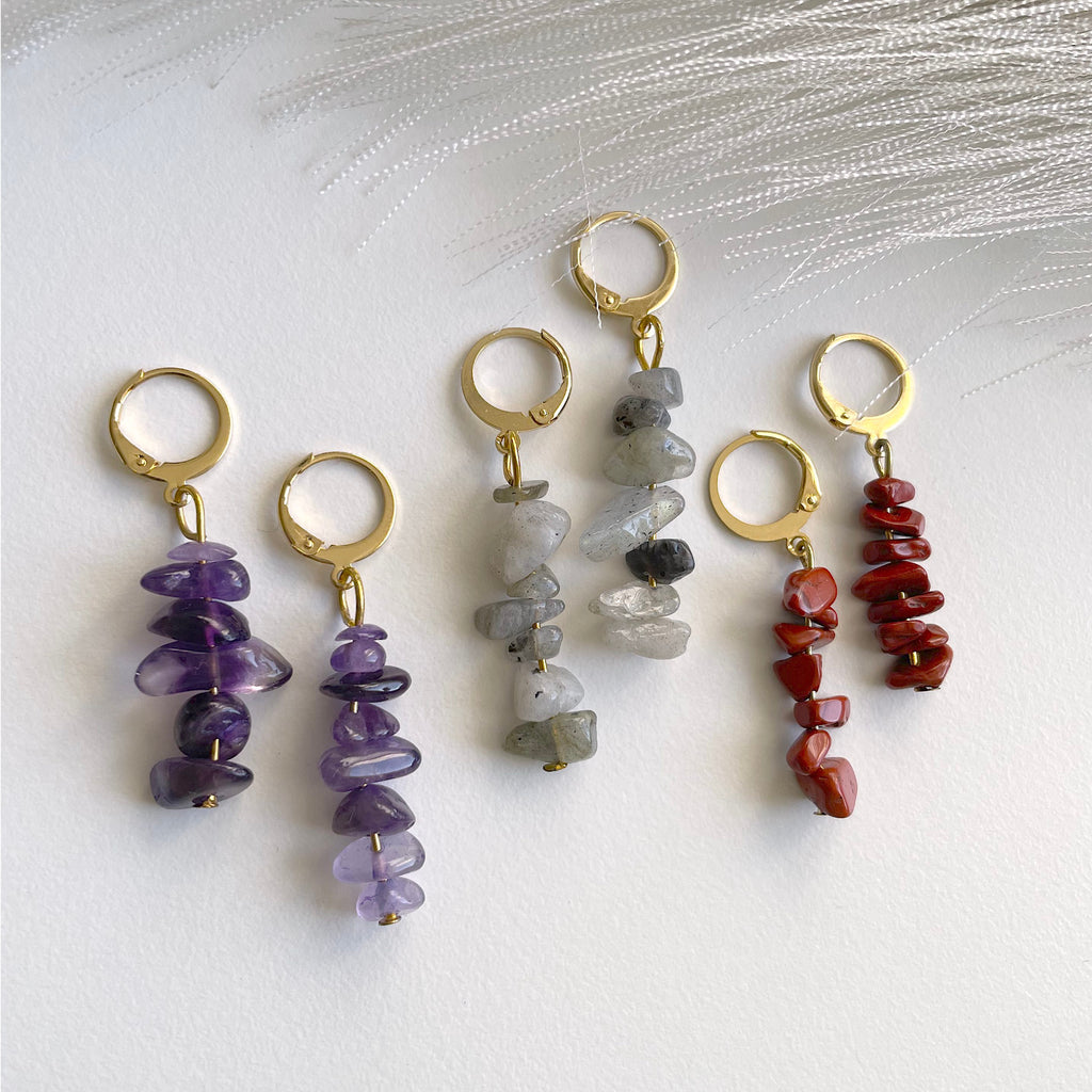 Three pairs of gold plated huggie hoop earrings with dangling irregular natural stone chips, arranged on a white surface with soft white feather-like decor. The stones are purple amethyst, gray quartz, and red jasper.