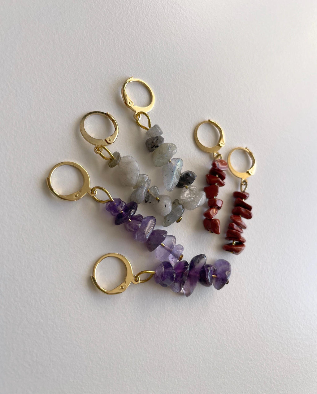 Four gold-tone earrings with dangling chip bead stones in purple amethyst, gray quartz, and red jasper, arranged on a plain off-white surface.