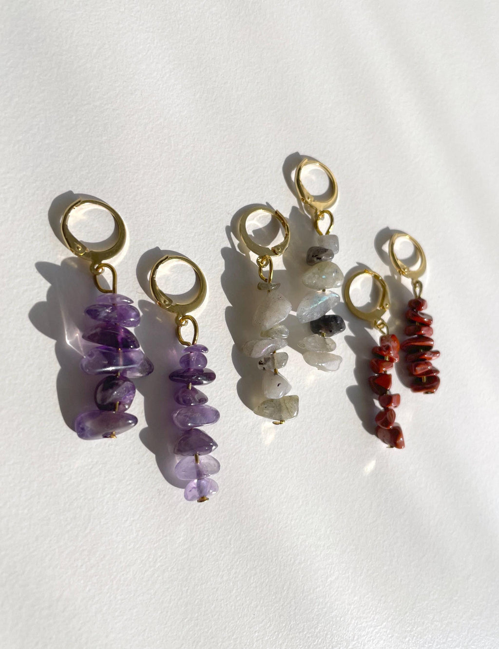 Three pairs of gold-tone hoop earrings with dangling stone charms, displayed on a white background. One pair features purple amethyst chips, one features milky white and gray quartz chips, and one features red jasper chips.