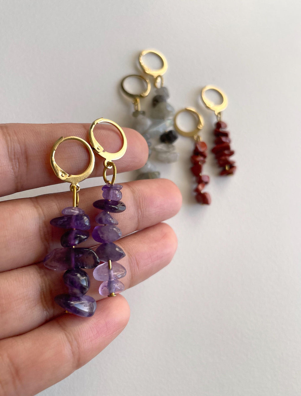 A close-up of a hand holding a pair of gold hoop earrings with dangling purple amethyst stone chip beads, with two additional pairs of earrings blurred in the background against a plain light wall.