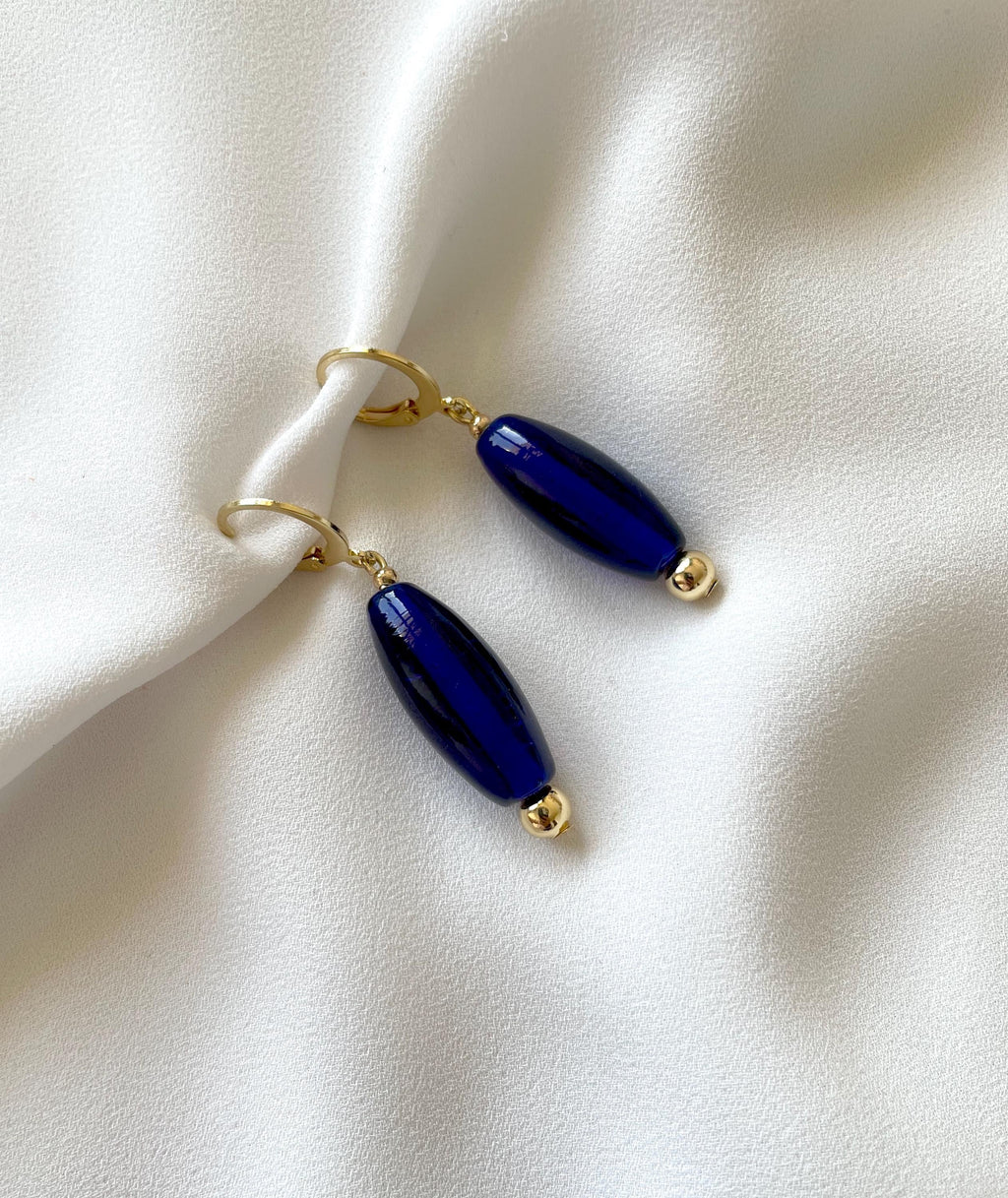 A pair of gold-tone drop earrings with glossy dark blue oval beads, displayed on a draped white fabric background.