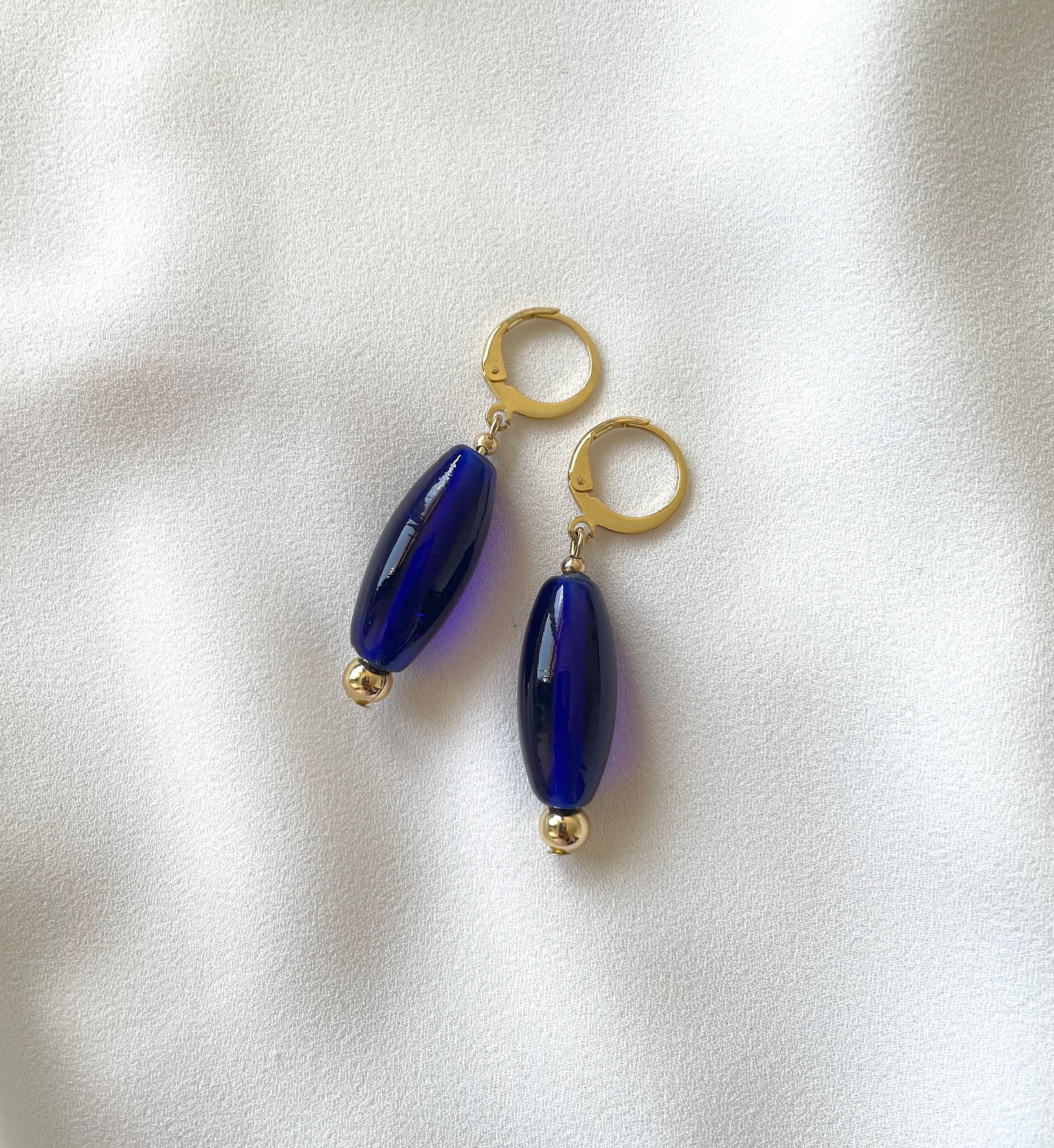 A pair of drop earrings with glossy dark blue oval beads and gold-tone hoops, displayed on a soft white fabric background.