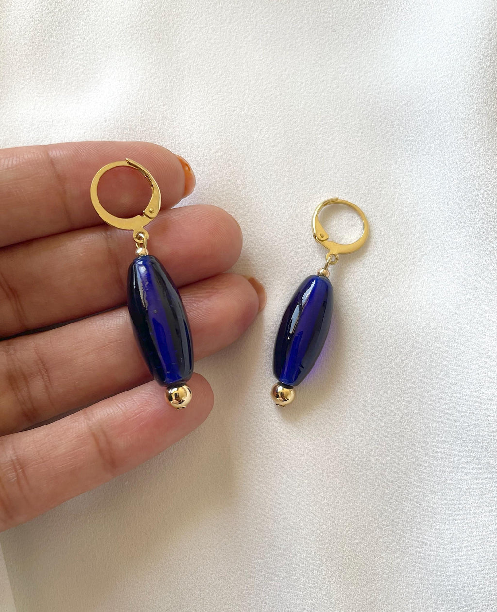 A pair of drop earrings with gold huggie hoops and glossy dark blue oval beads, held between fingers against a white fabric background.