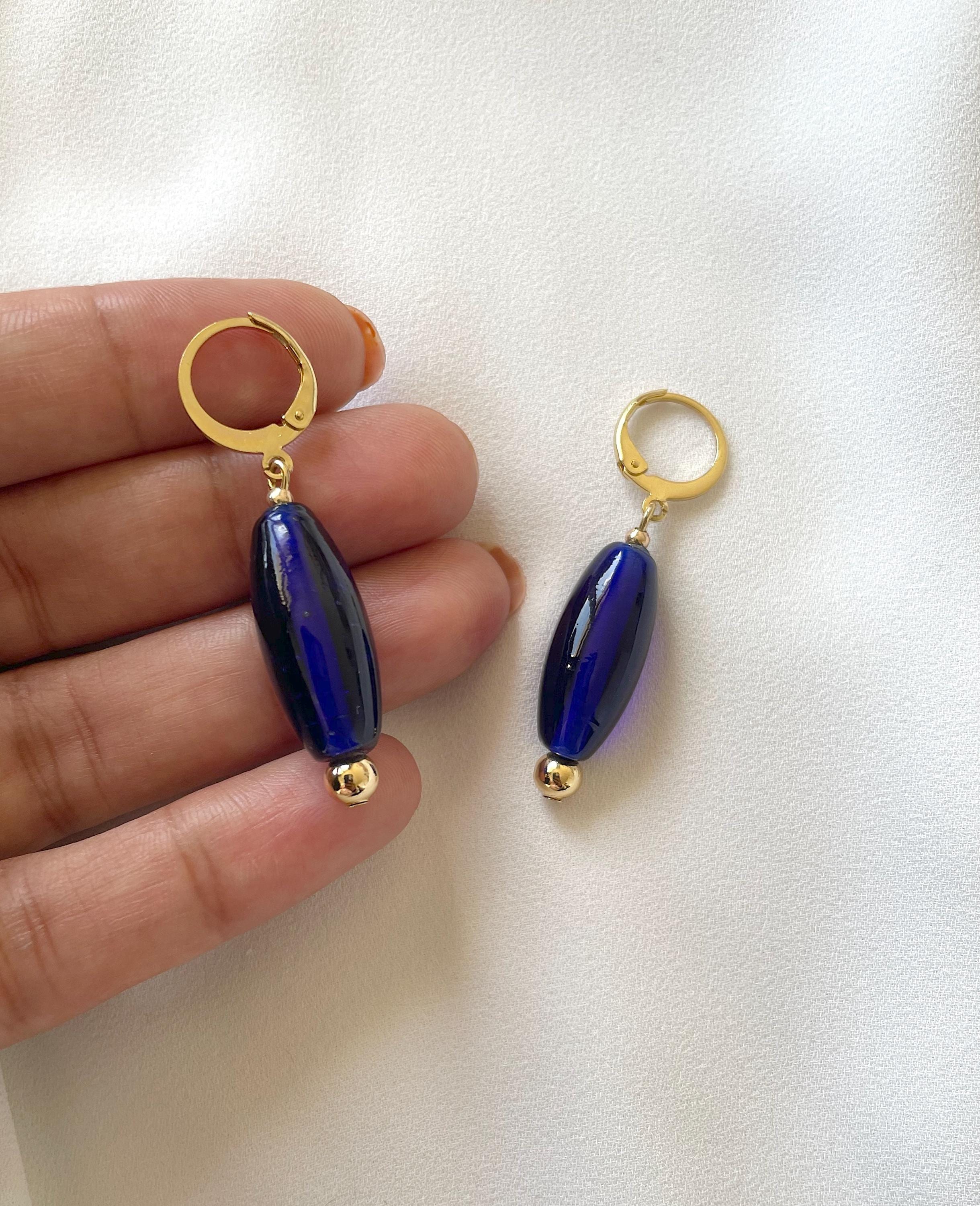 A pair of drop earrings with gold huggie hoops and glossy dark blue oval beads, held between fingers against a white fabric background.