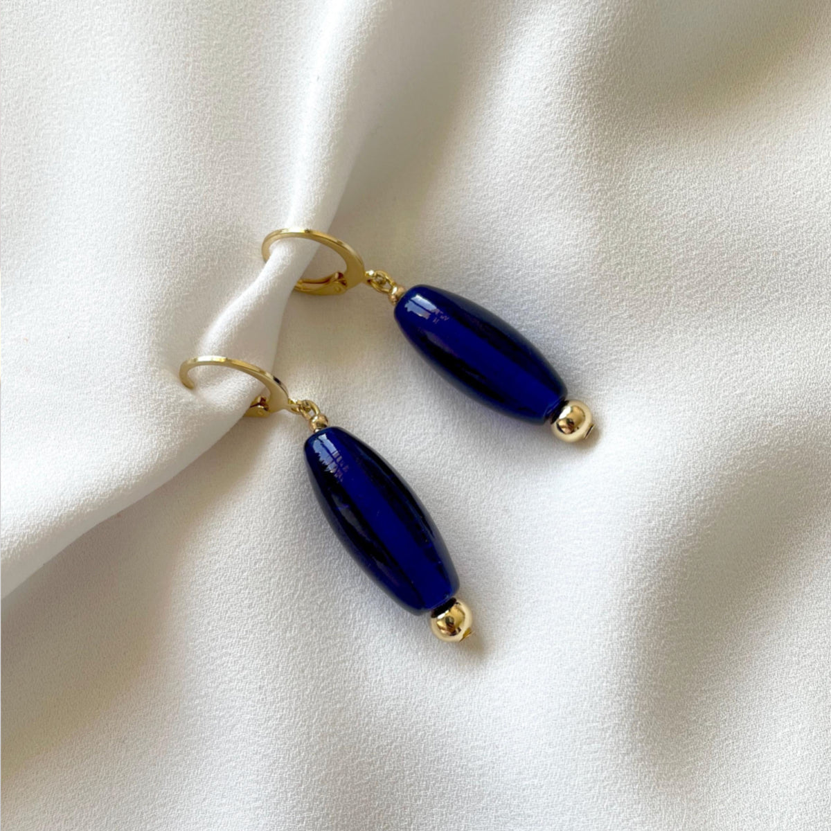 A pair of gold-tone 18K gold plated drop earrings with glossy navy blue oval glass beads followed by gold toned metal beads, attached to huggie hoops, displayed on a draped white fabric background.