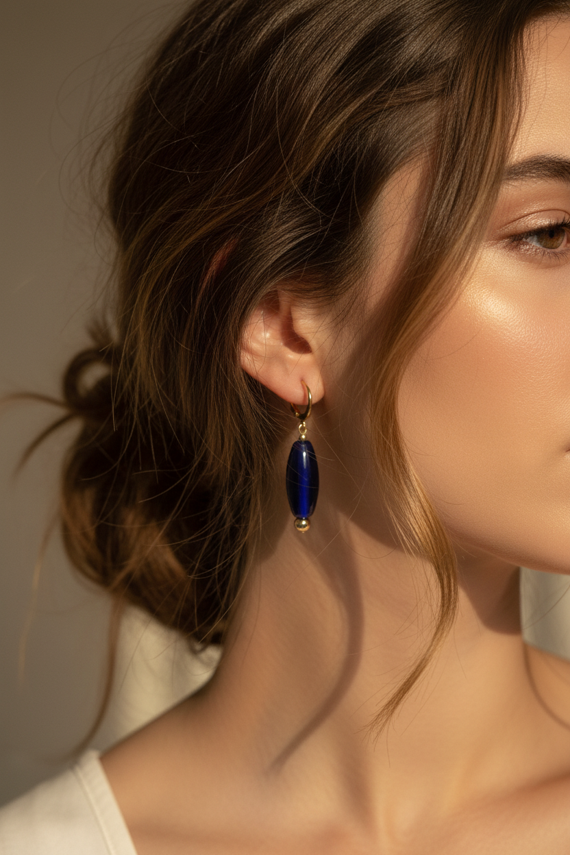 A close-up of a woman wearing a single drop earring with a polished dark blue oval stone set in a gold-tone huggie hoops.