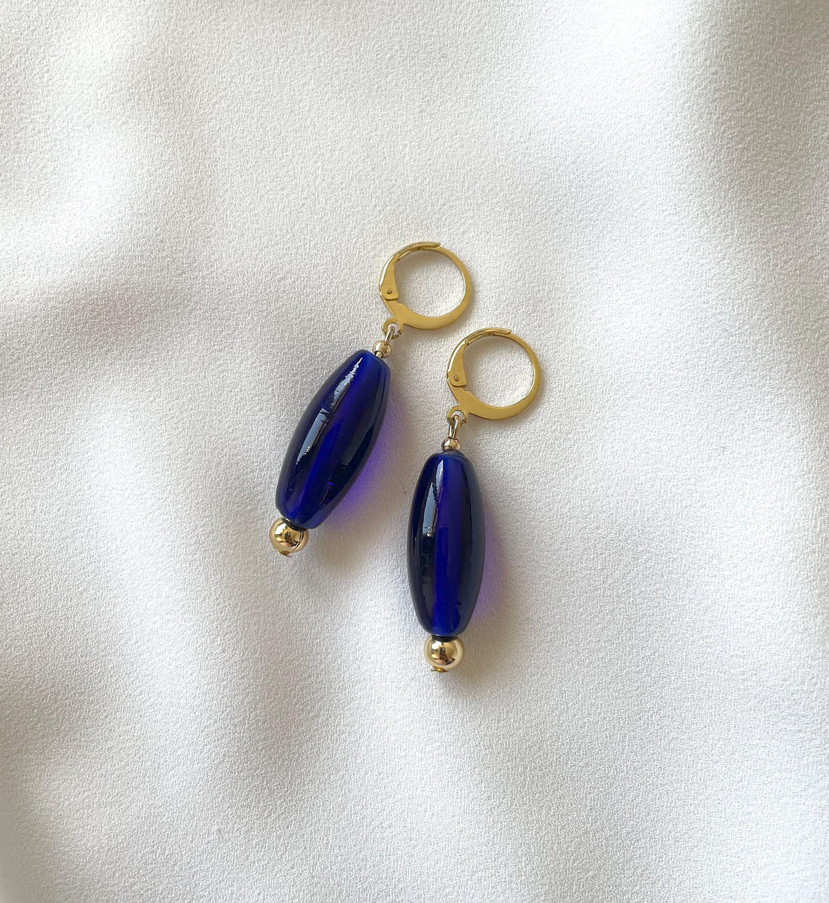 A pair of drop earrings with glossy dark blue oval beads and gold-tone hoops, displayed on a soft white fabric background.