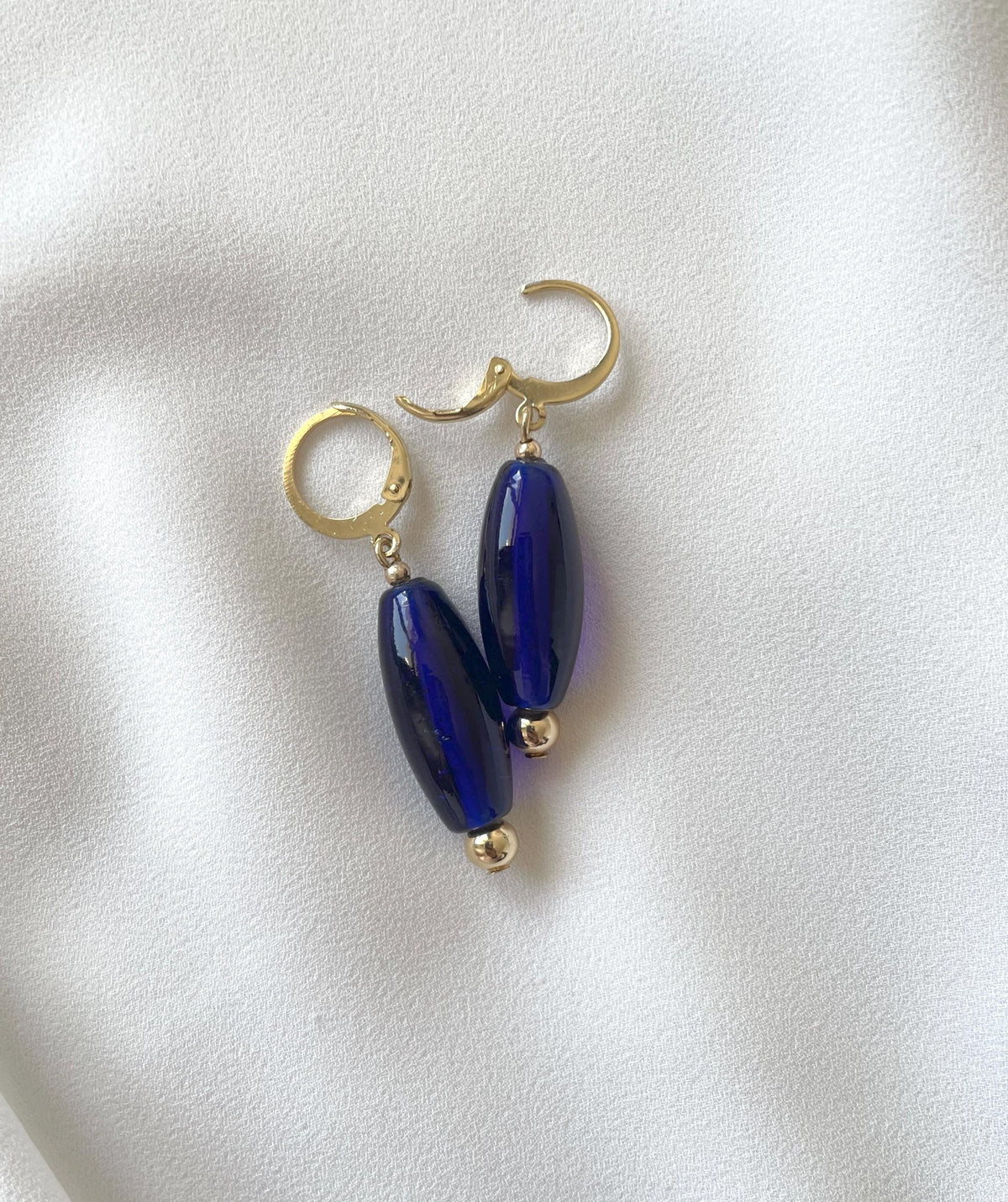 A pair of gold hoop earrings with elongated, glossy dark blue glass beads, displayed on a soft, white textured fabric background.