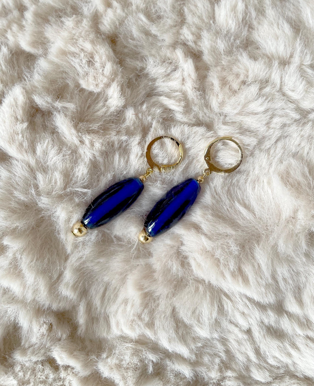 A pair of drop earrings with glossy navy blue glass beads and gold-tone hoops, displayed on a white faux fur surface.