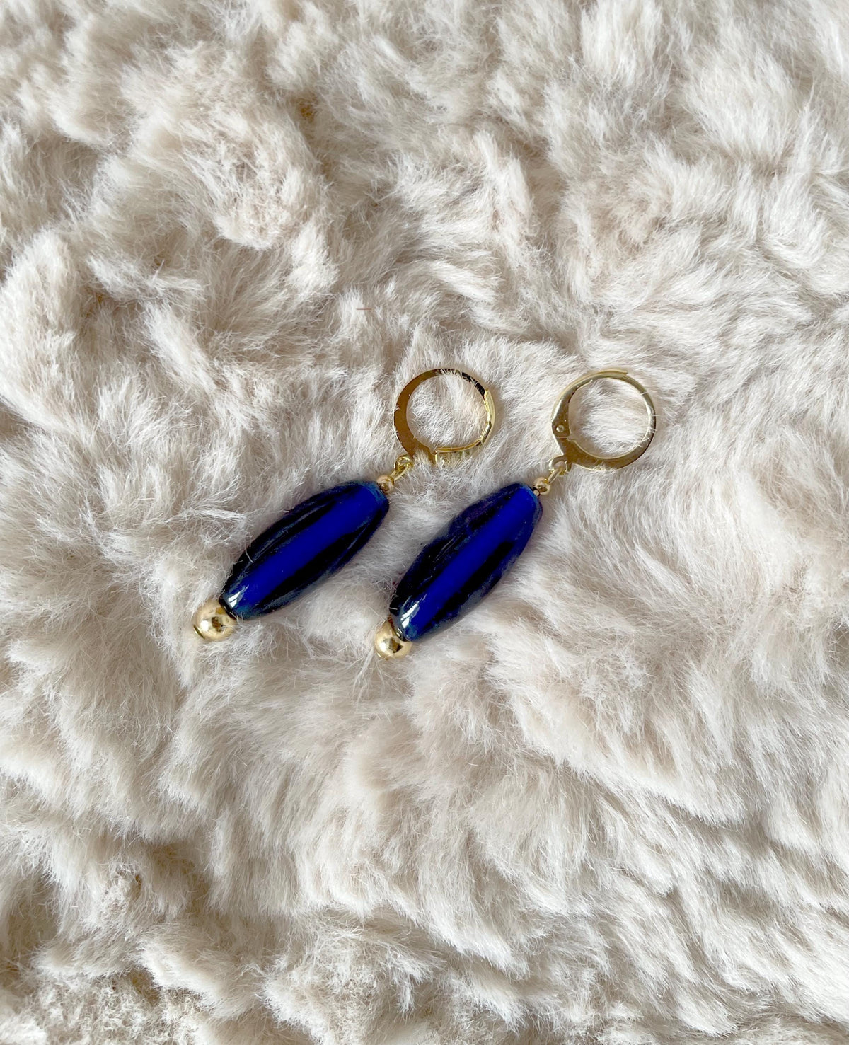 A pair of drop earrings with glossy navy blue glass beads and gold-tone hoops, displayed on a white faux fur surface.