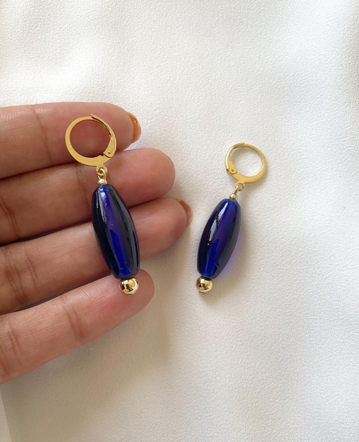 A pair of drop earrings with gold huggie hoops and glossy navy blue oval beads, held between fingers against a white fabric background.