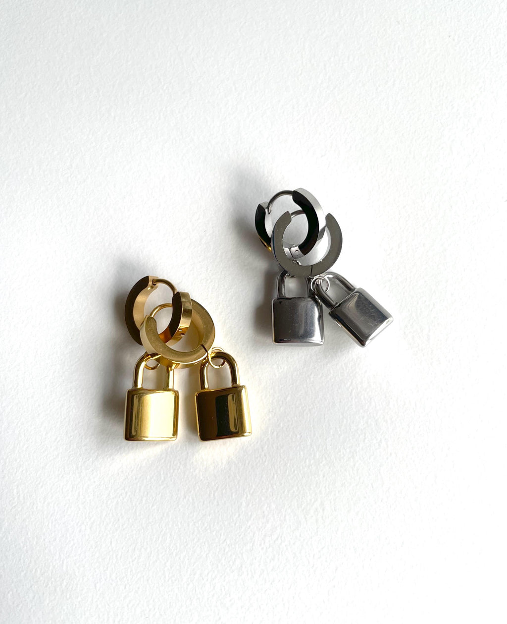 Two pairs of small hoop earrings with dangling padlock charms, one in polished gold and one in polished silver, displayed on a clean white background.