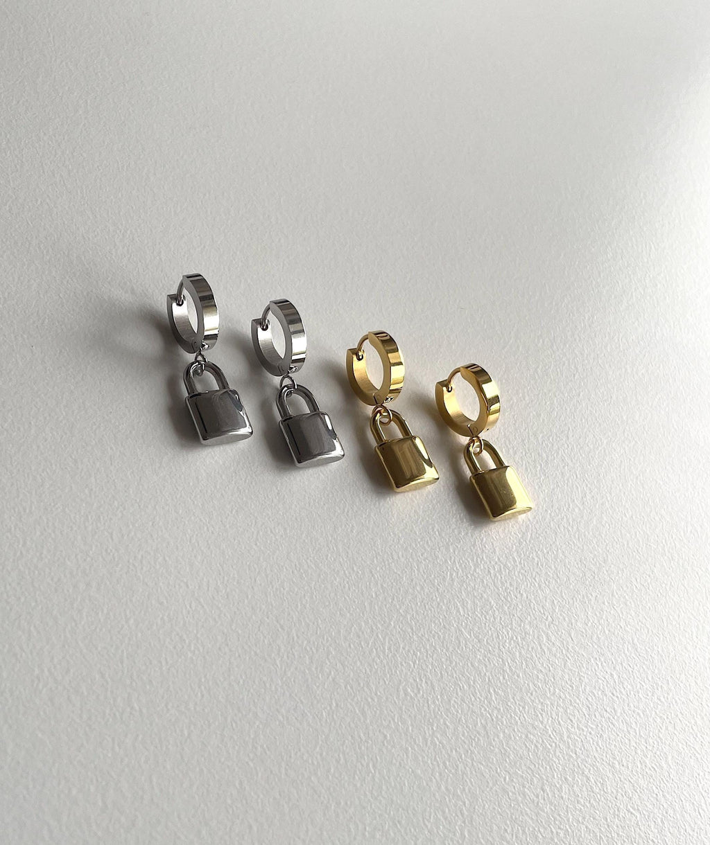 Four small hoop earrings with dangling padlock charms, shown in two metal finishes: silver and gold. Two silver pairs are on the left and two gold pairs are on the right, arranged on a light gray textured surface.