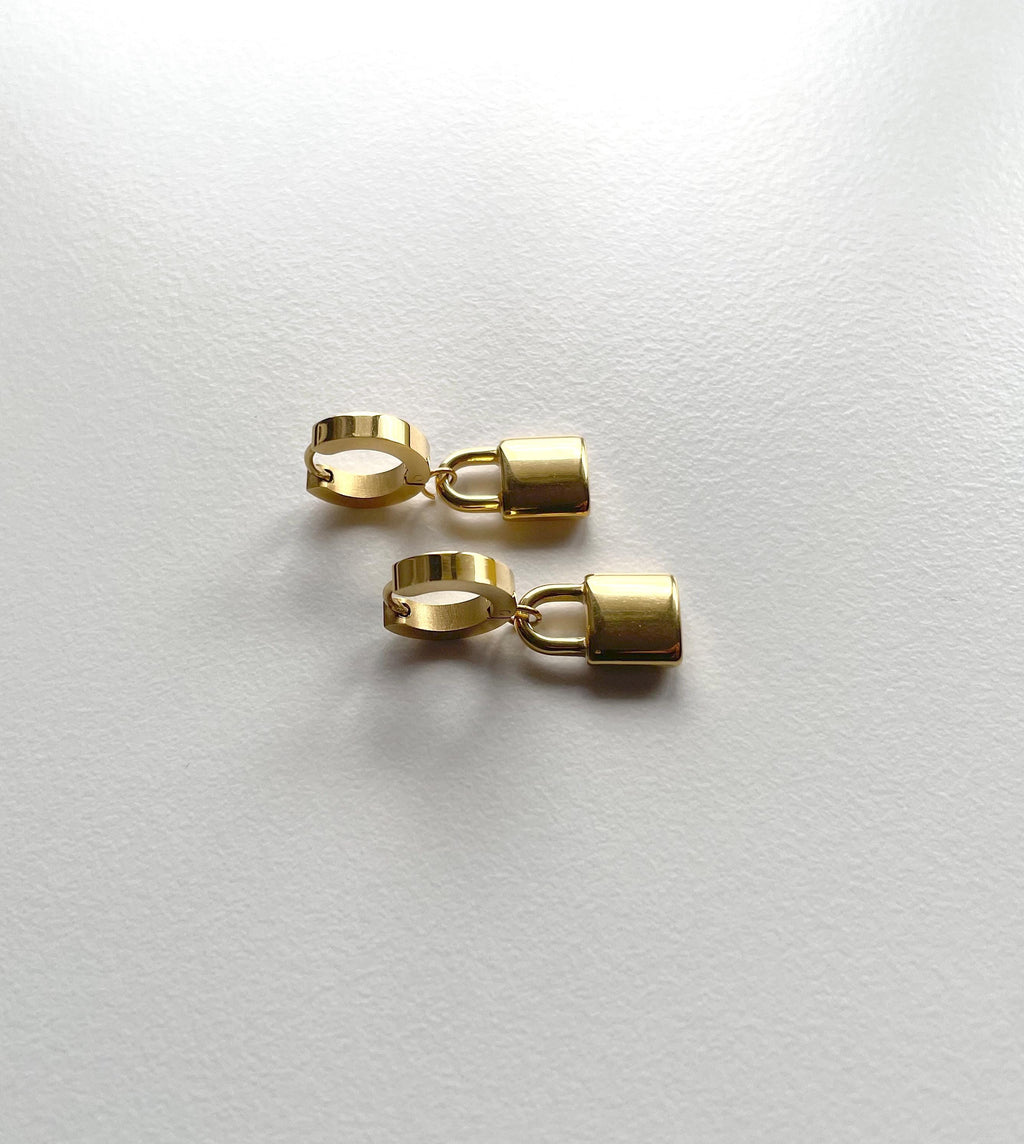 A pair of gold-toned huggie hoop earrings with small rectangular pendant charms, displayed on a clean white textured surface.