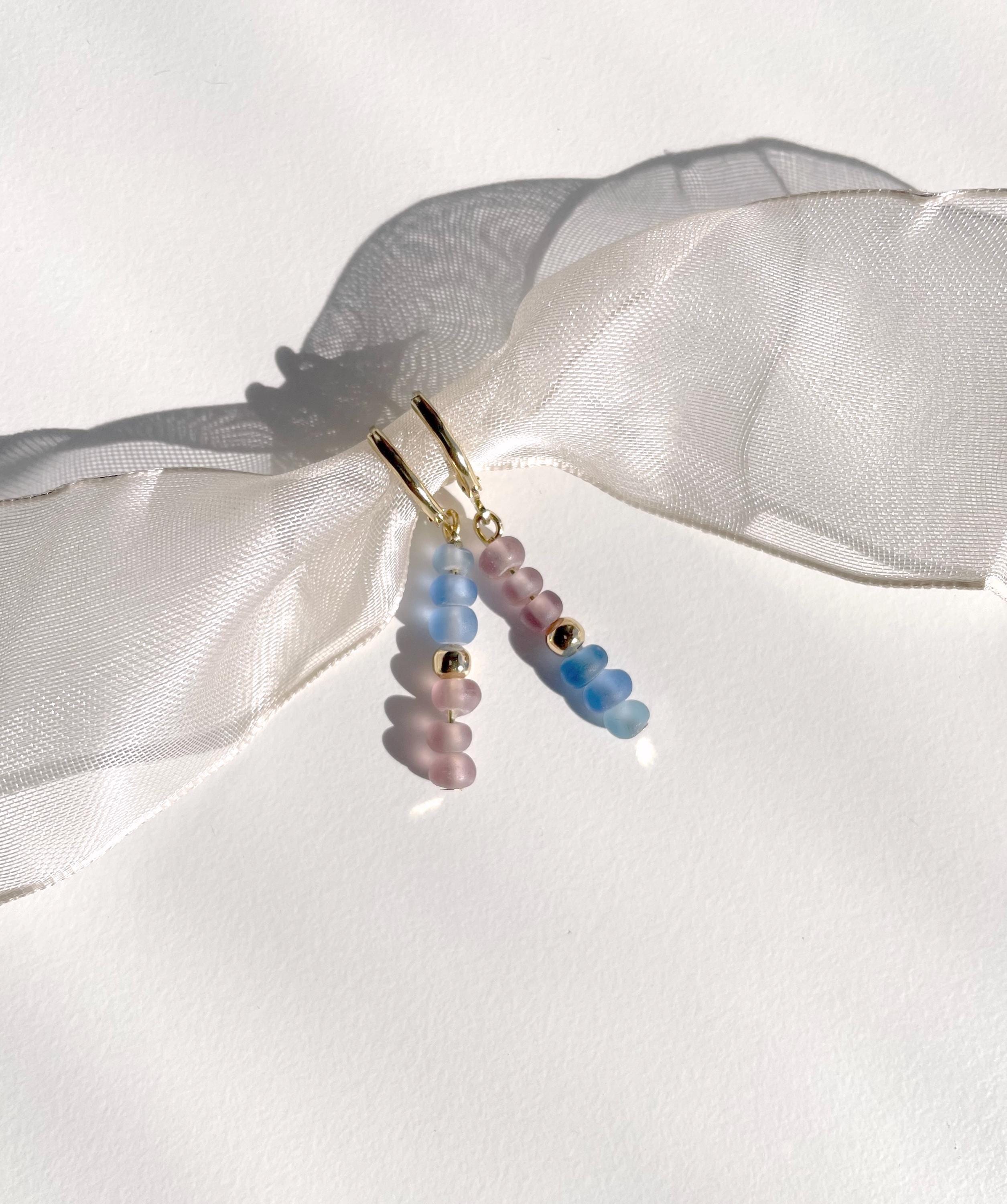 A pair of dangling beaded earrings with gold-tone hooks, featuring translucent pastel beads in pink, blue, and lavender, displayed against a white background with a sheer white ribbon.