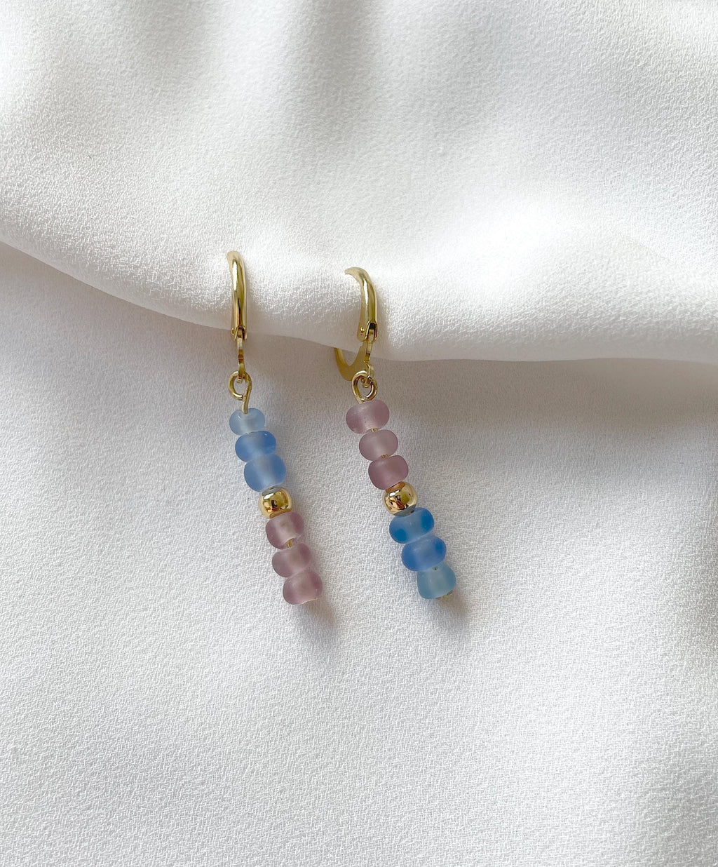A pair of gold huggie hoop earrings with dangling beaded chains featuring light blue and pink round beads, displayed on a soft white fabric background.