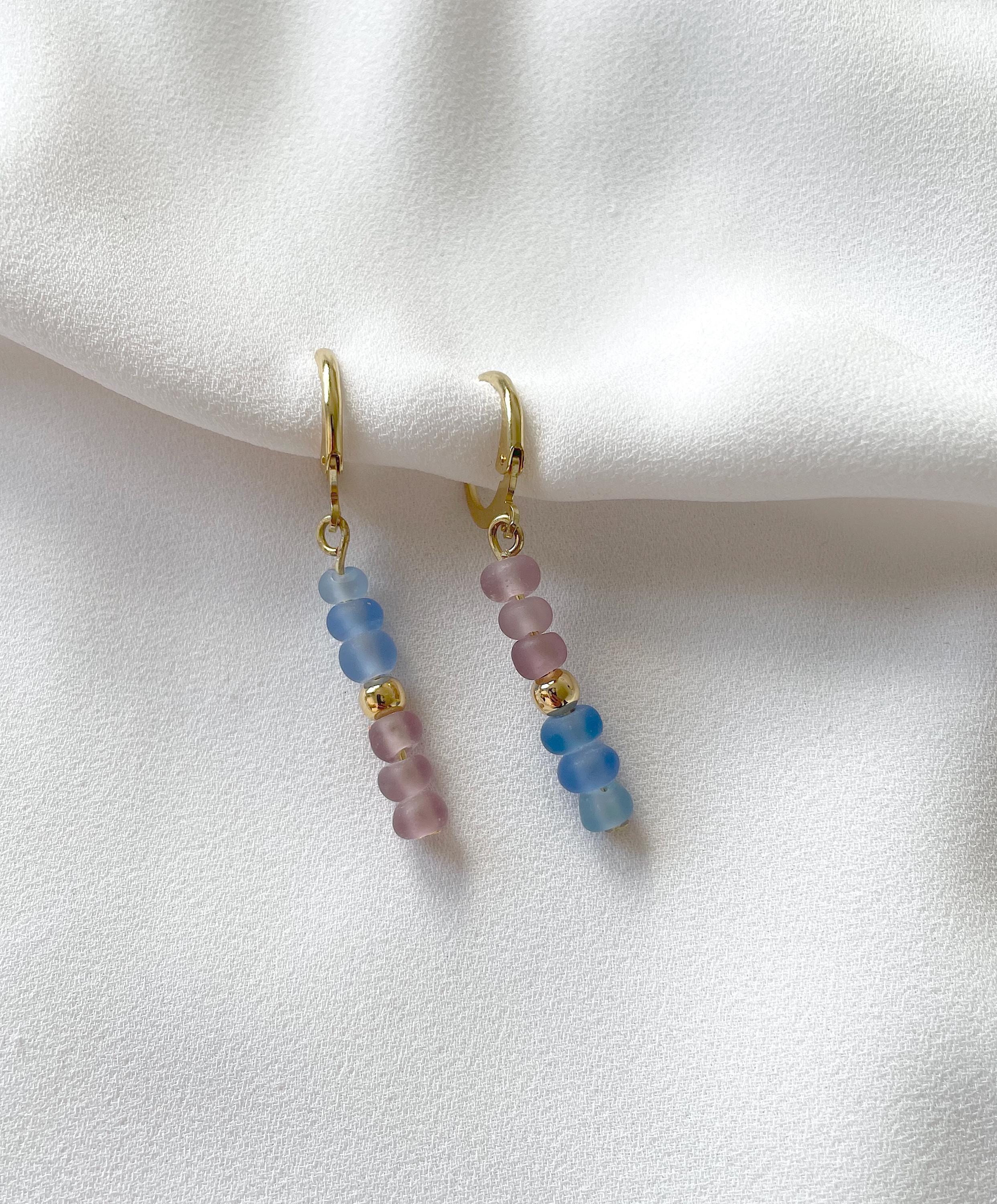 A pair of gold huggie hoop earrings with dangling beaded chains featuring light blue and pink round beads, displayed on a soft white fabric background.