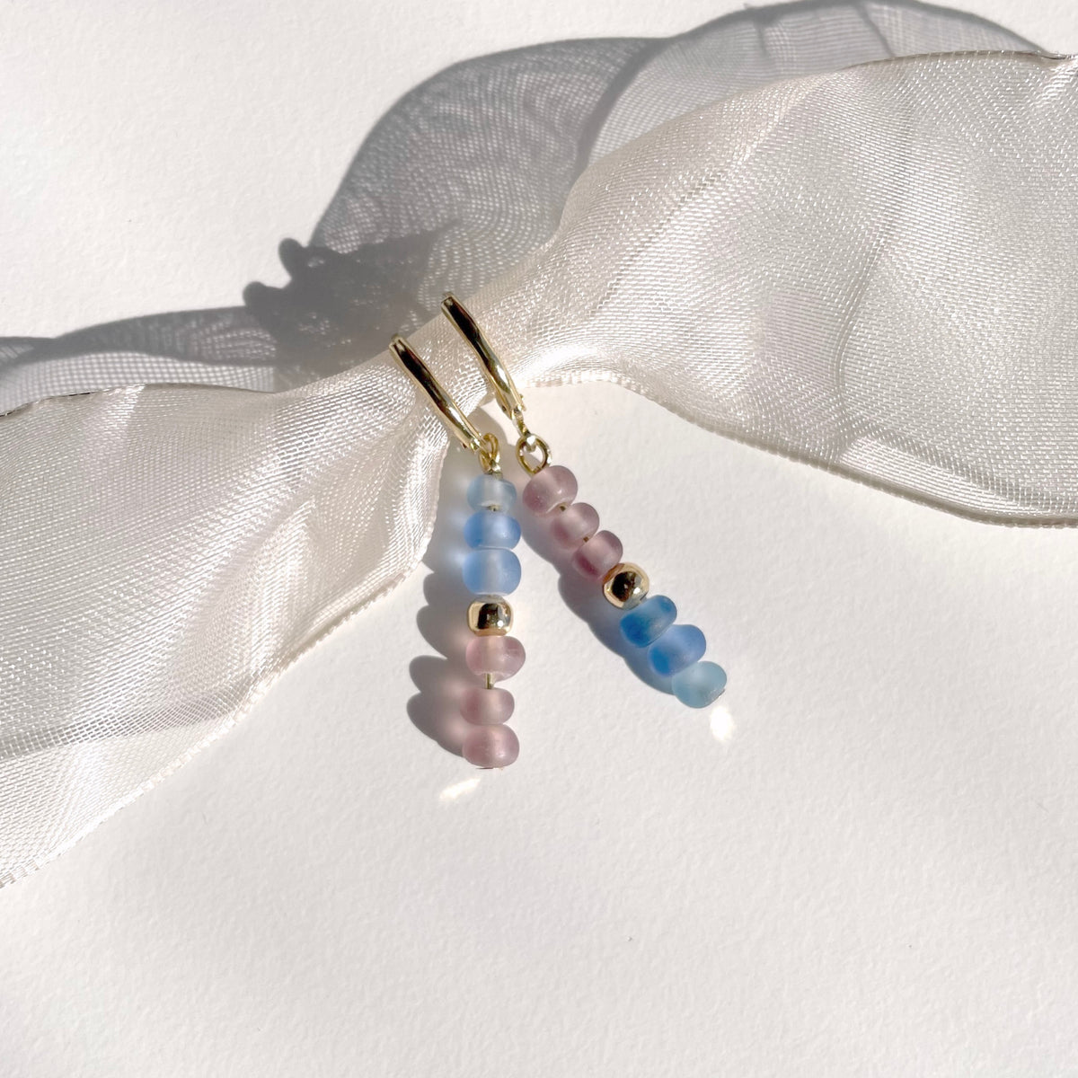 A pair of dangling beaded earrings with gold-tone huggie hoops, featuring translucent pastel beads in pink, blue, and gold, displayed against a white background with a sheer white ribbon.