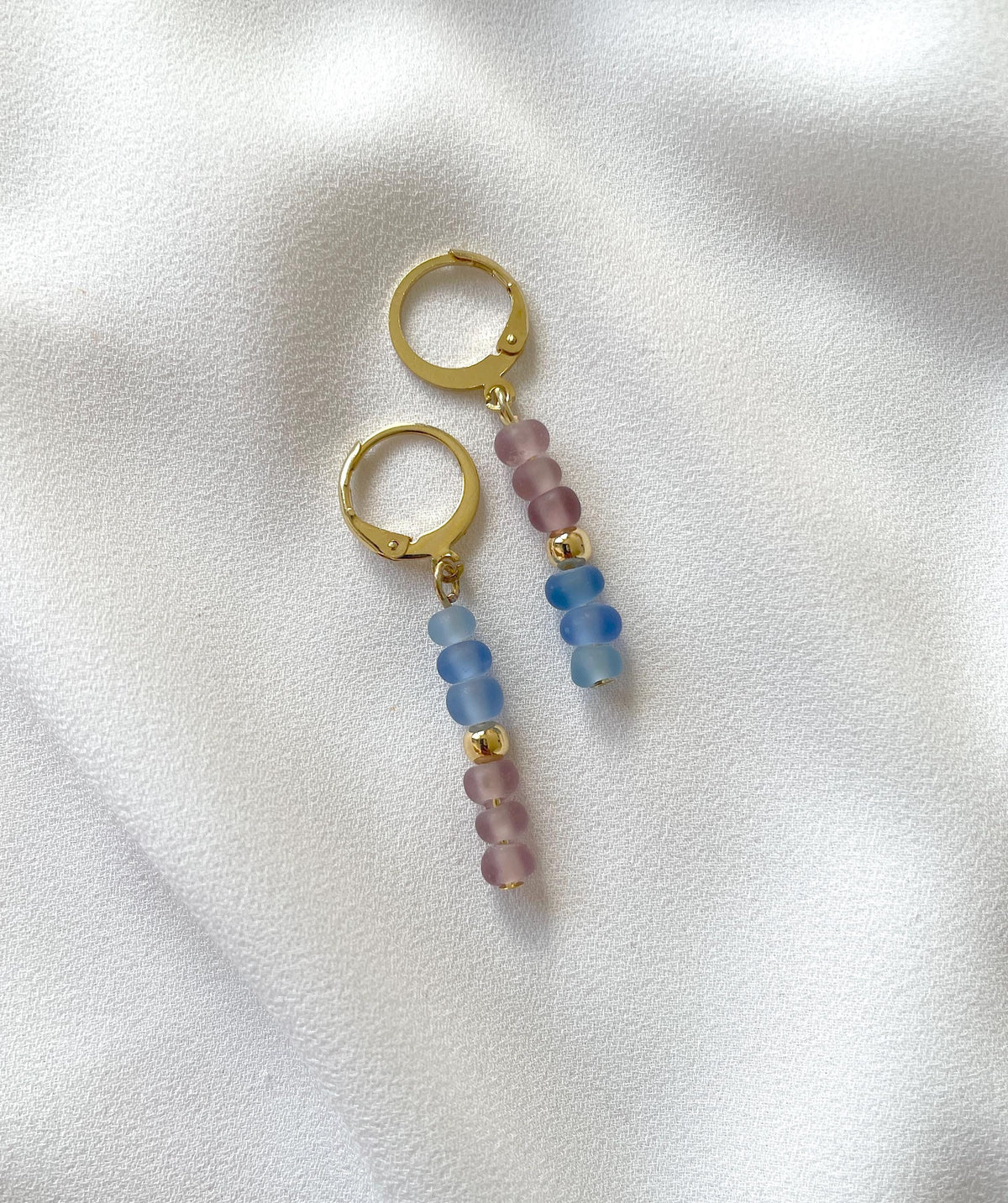 A pair of golden huggie hoop dangle earrings with translucent beads in shades of powder blue and mauve pink, resting on a textured white fabric background.