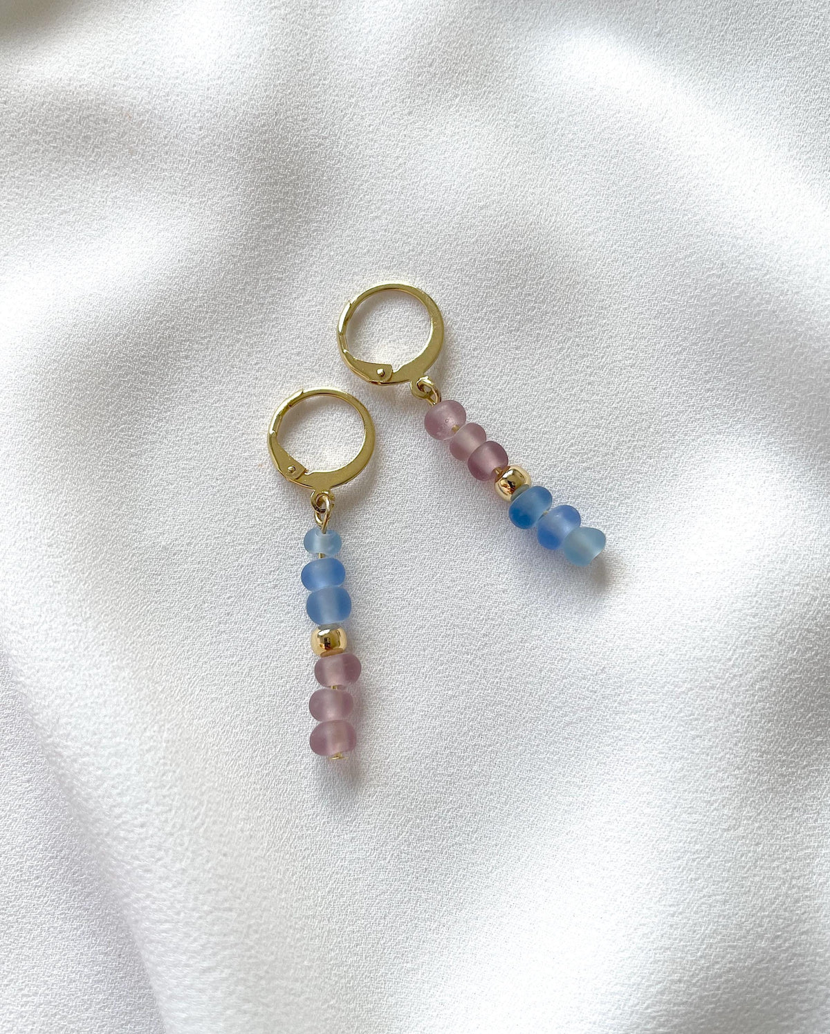A pair of golden huggie hoop earrings with dangling beaded chains featuring pastel light blue and dusty pink beads, displayed on a soft white fabric background.