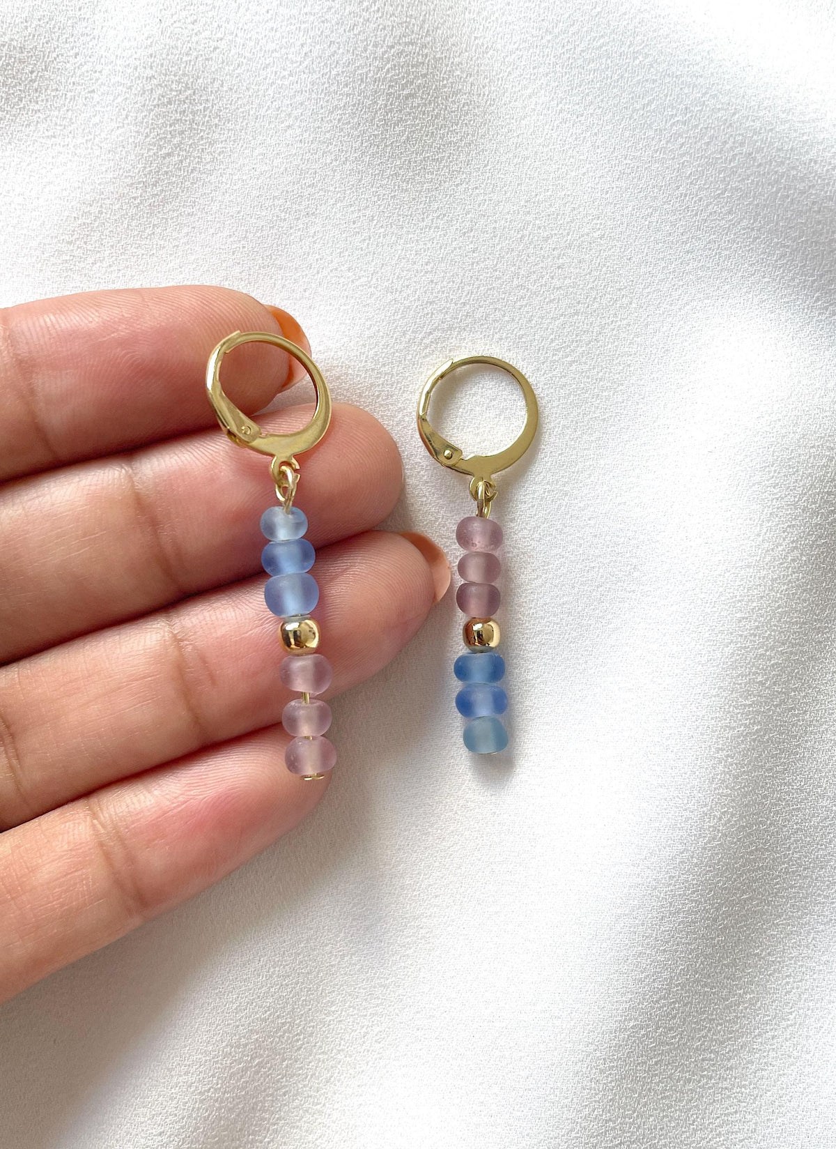 A pair of gold plated huggie hoop dangle earrings with translucent pastel blue and pink beads, held by a hand against a white textured fabric background.