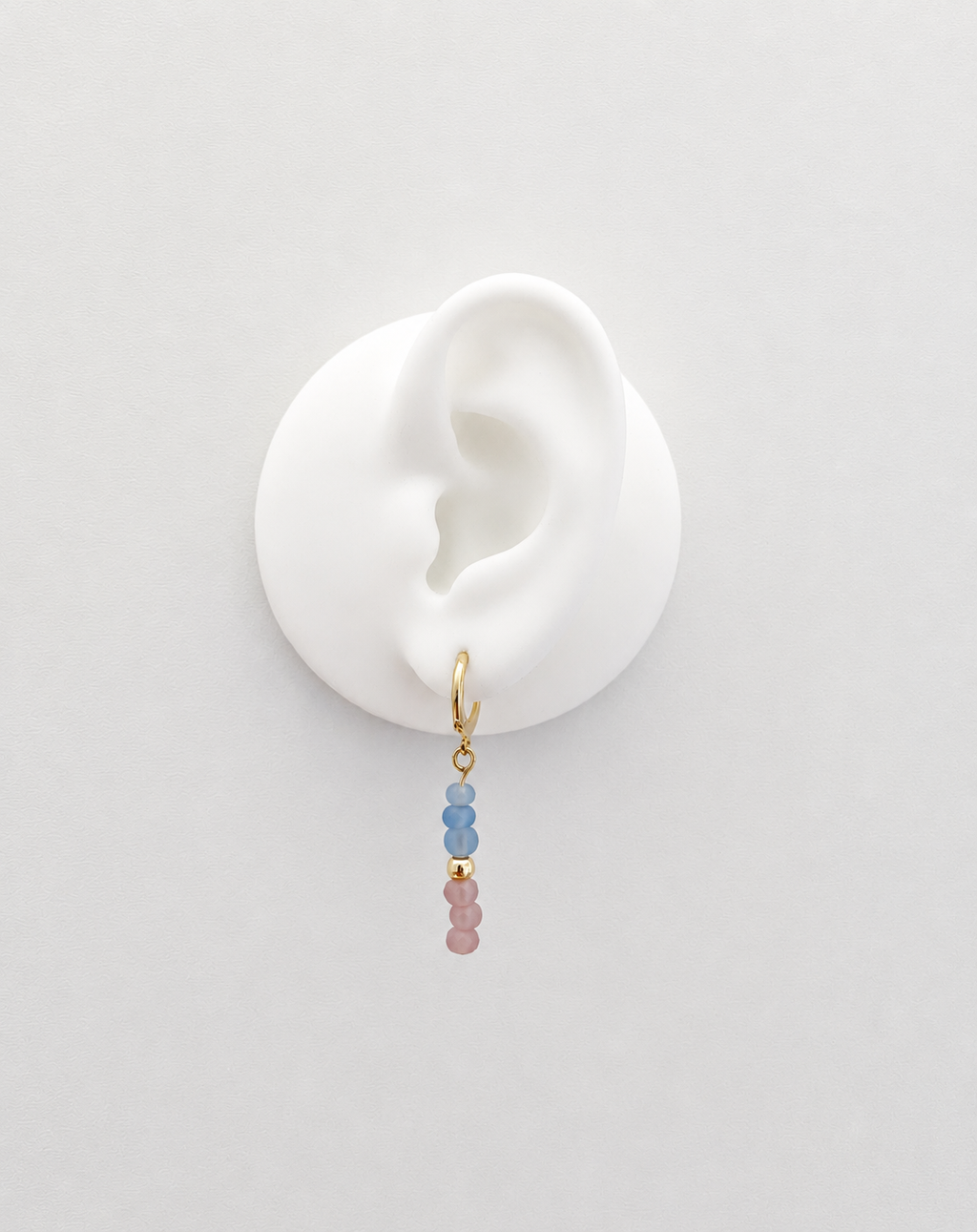 A delicate gold hoop earring with three small beads in light blue and dusty pink, displayed on a white silicone ear stand against a light gray background.