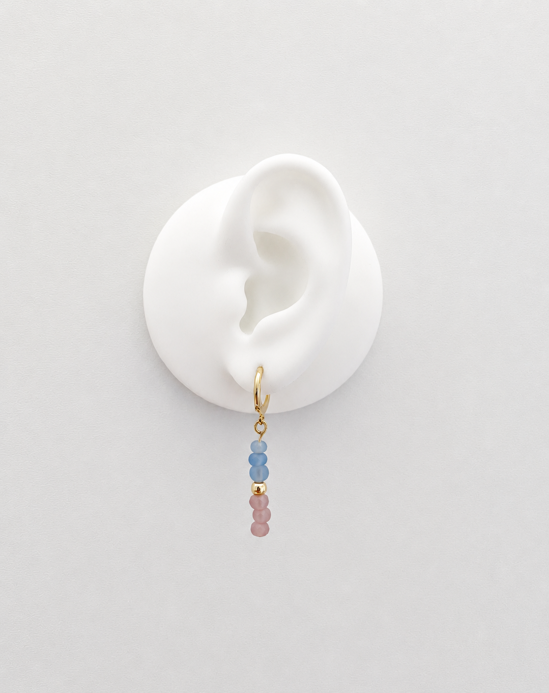 A delicate gold hoop earring with three small beads in light blue and dusty pink, displayed on a white silicone ear stand against a light gray background.