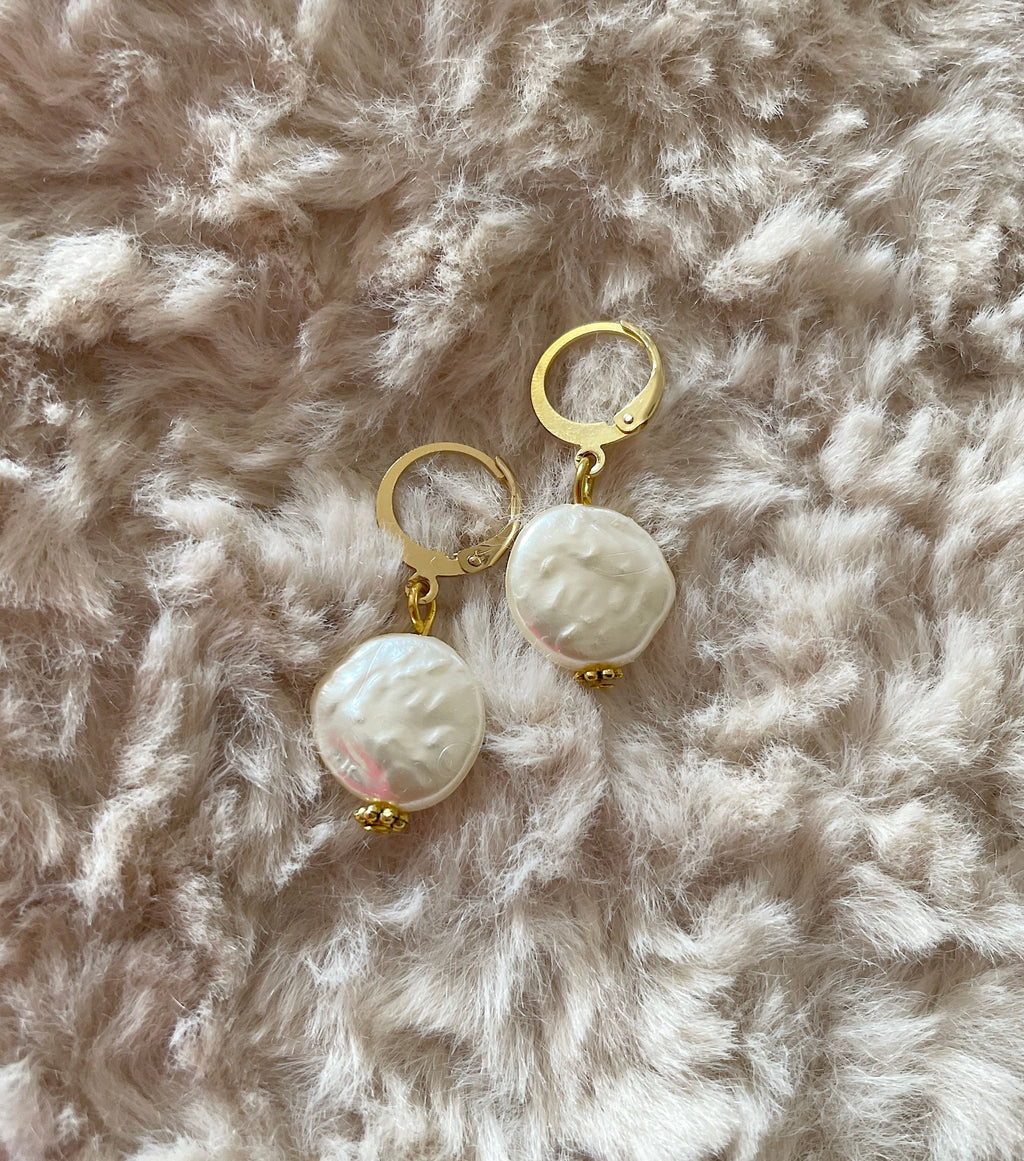A pair of gold plated pearl hoop earrings with large, irregular white faux baroque pearls, displayed on a soft, textured beige faux fur surface.