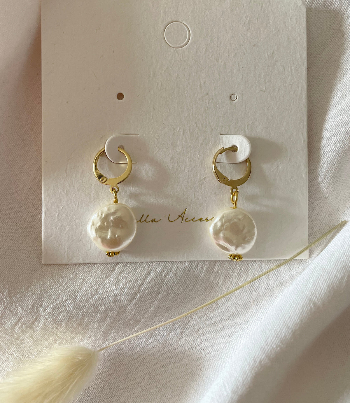 A pair of pearl huggie hoop earrings with large, textured white pearl drops, displayed on a white branded card labeled Kapella Accessories against a soft white fabric background.