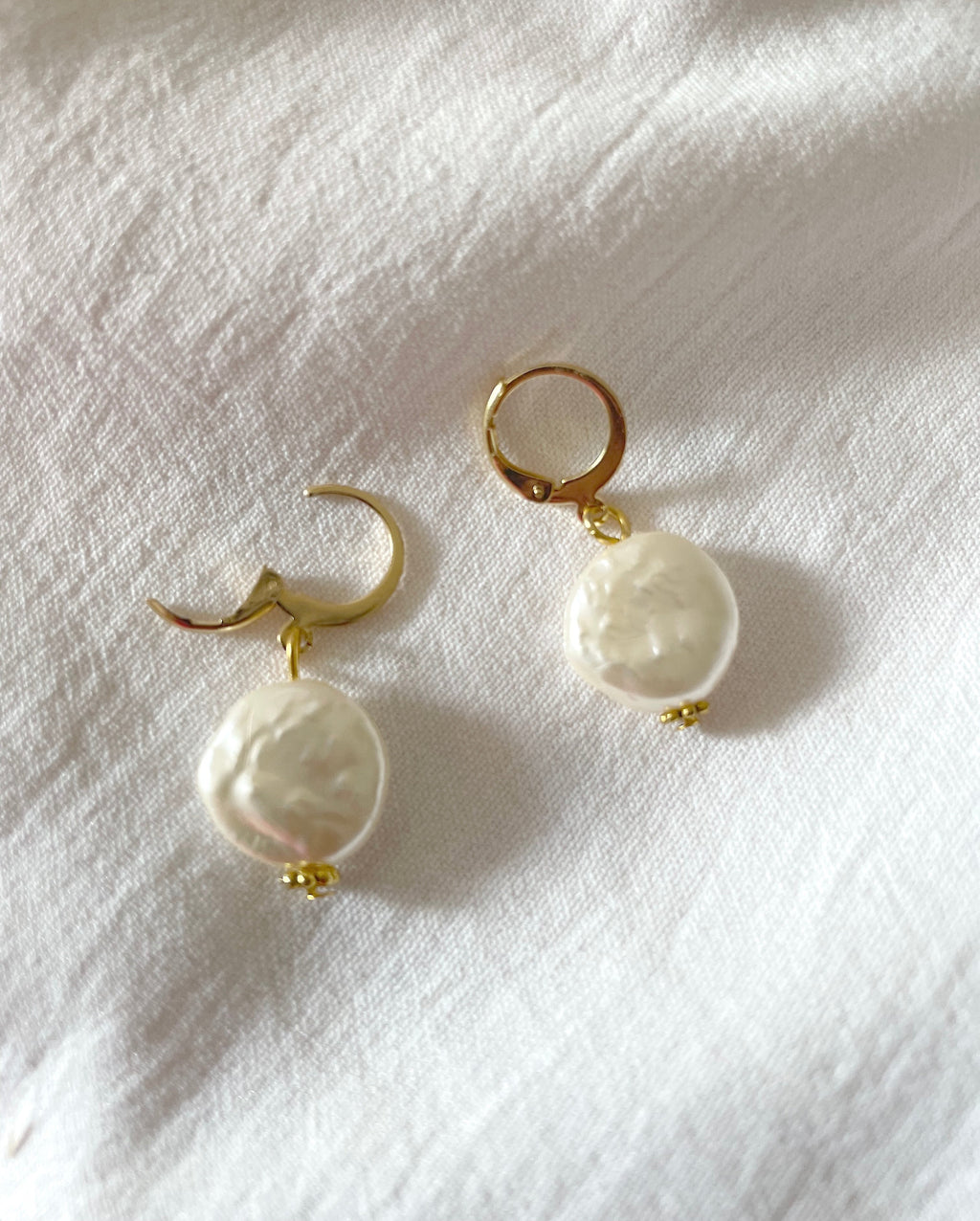 A pair of pearl huggie hoop earrings with large, irregular white baroque pearls hanging from them, displayed on a textured white fabric background.