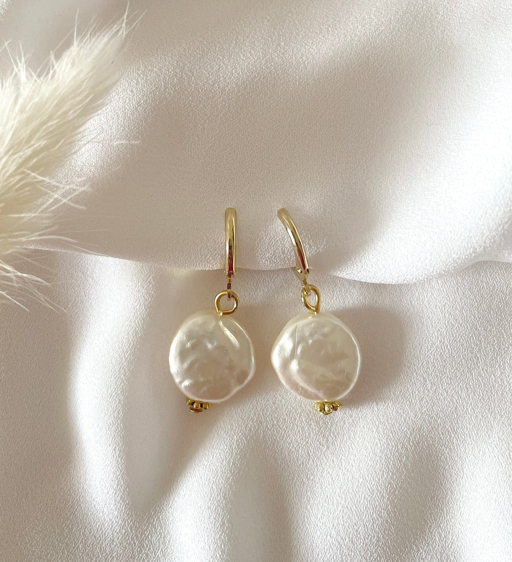 A pair of gold plated Huggies with irregular white baroque pearls, displayed on a soft white fabric background with a decorative beige feather.